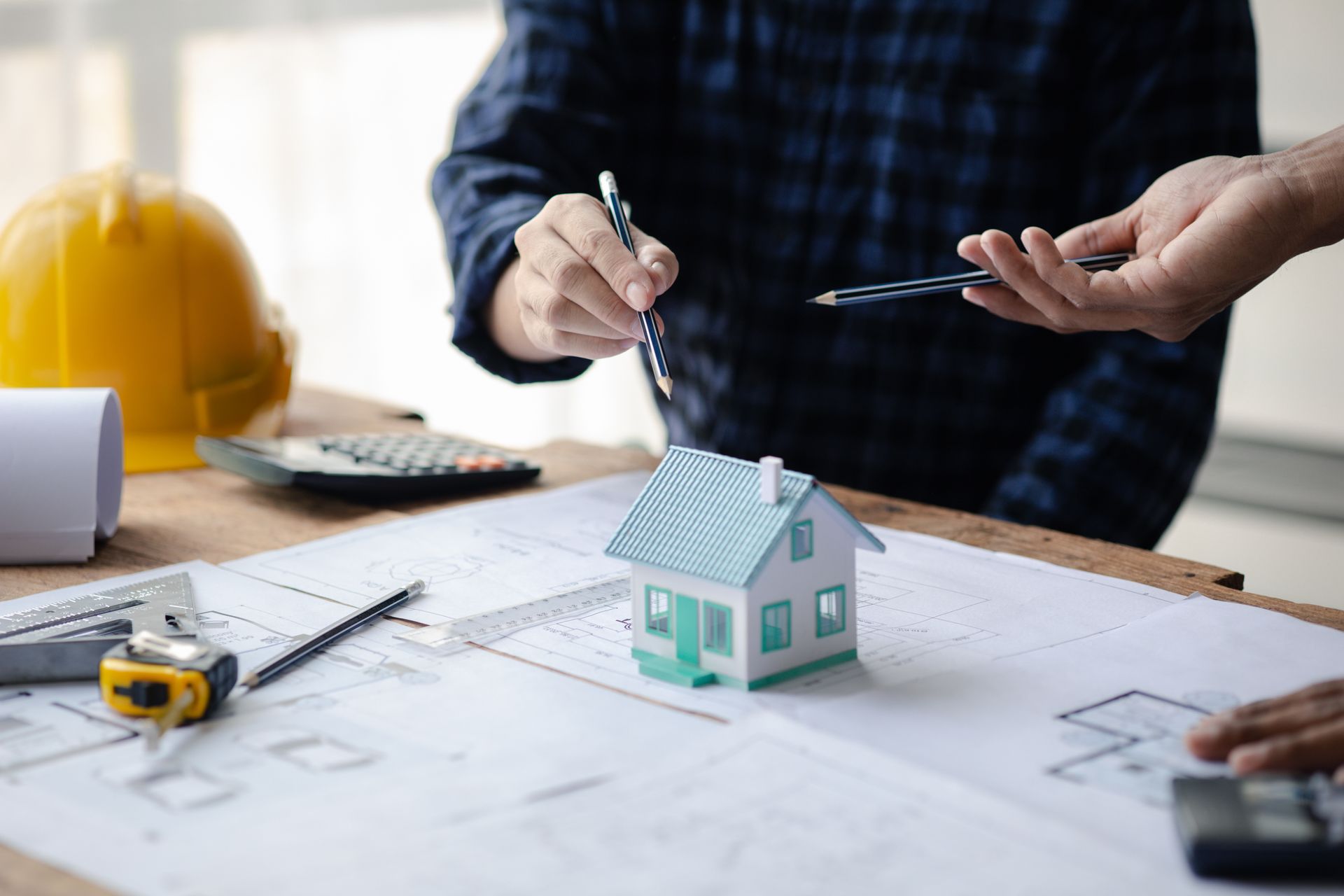 Hands drafting house plans beside a small model home on a desk with a yellow hard hat Hands drafting house plans beside a small model home on a desk with a yellow hard hat