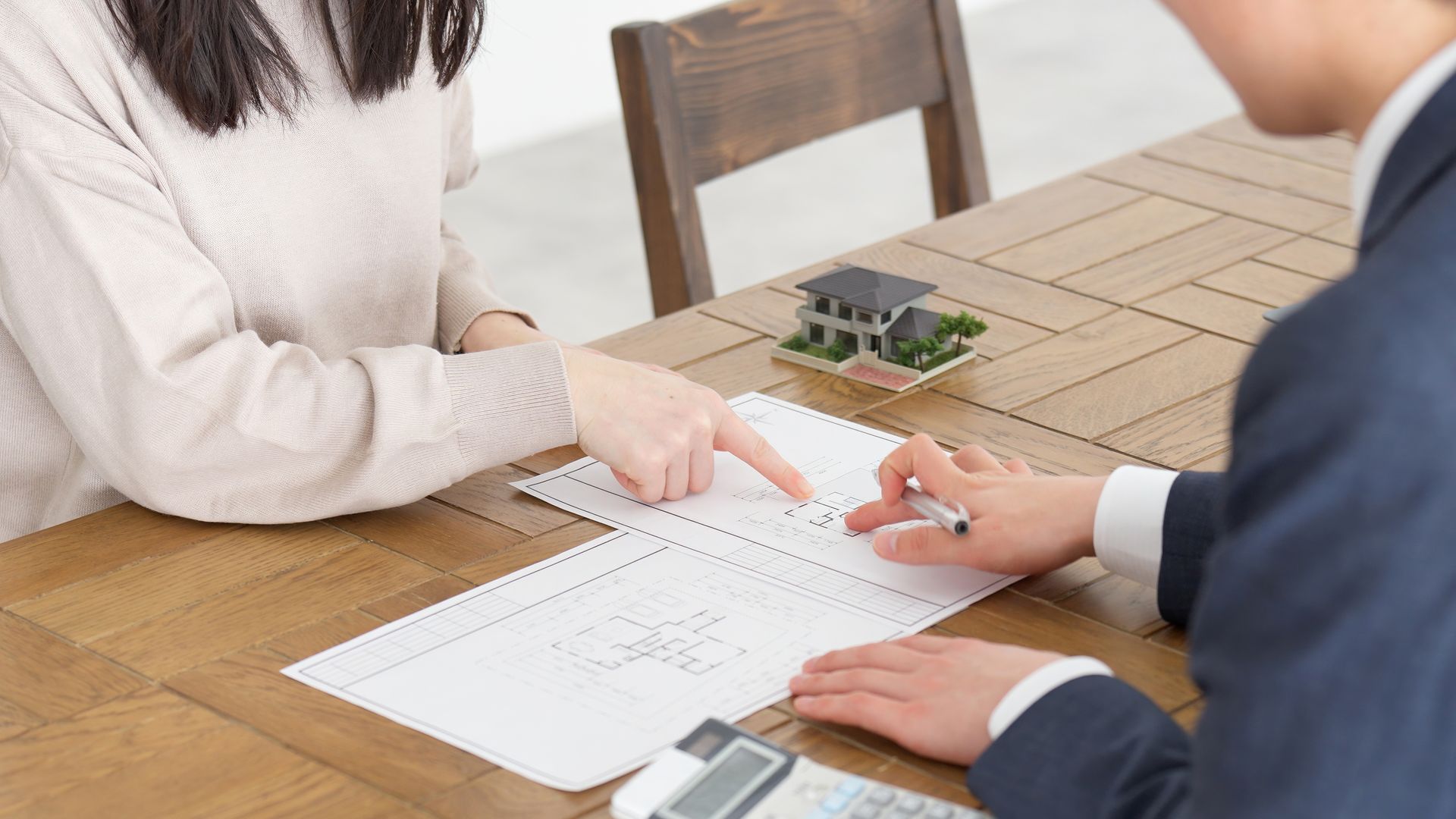 Two people review a floor plan at a wooden table, pointing at blueprints beside a small house model.