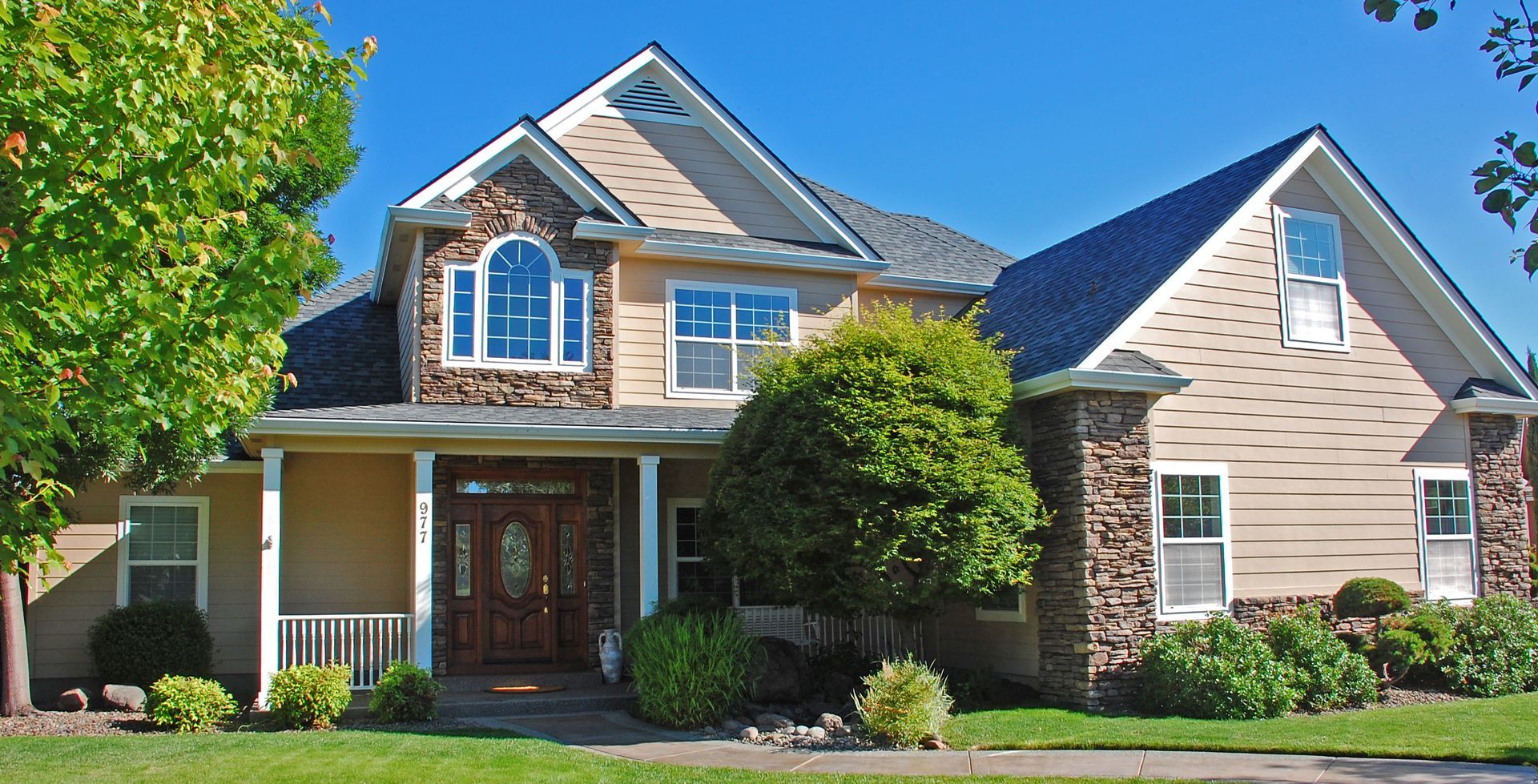 Suburban two-story house with beige siding, dark roof, front porch, and manicured lawn under a bright blue sky