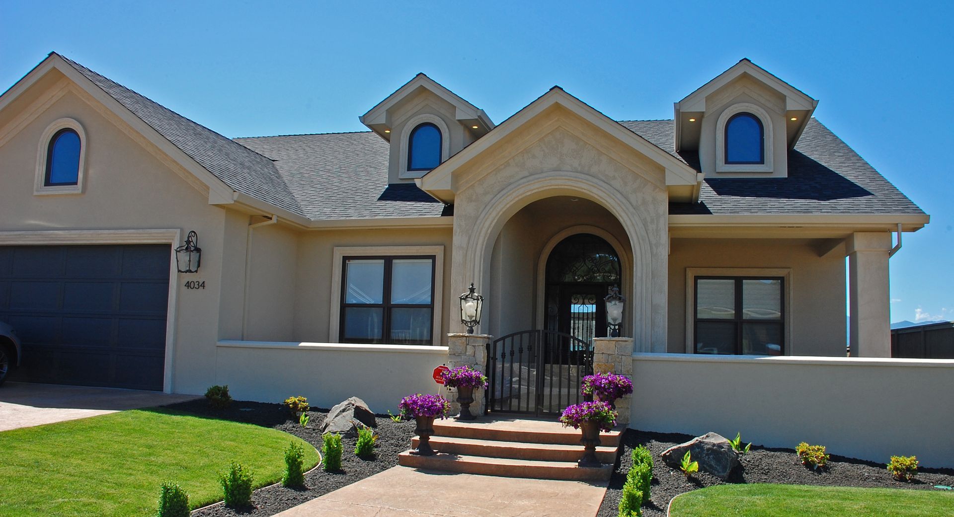 Single-story beige house with arched entry, blue dormers, and flower-lined front steps.
