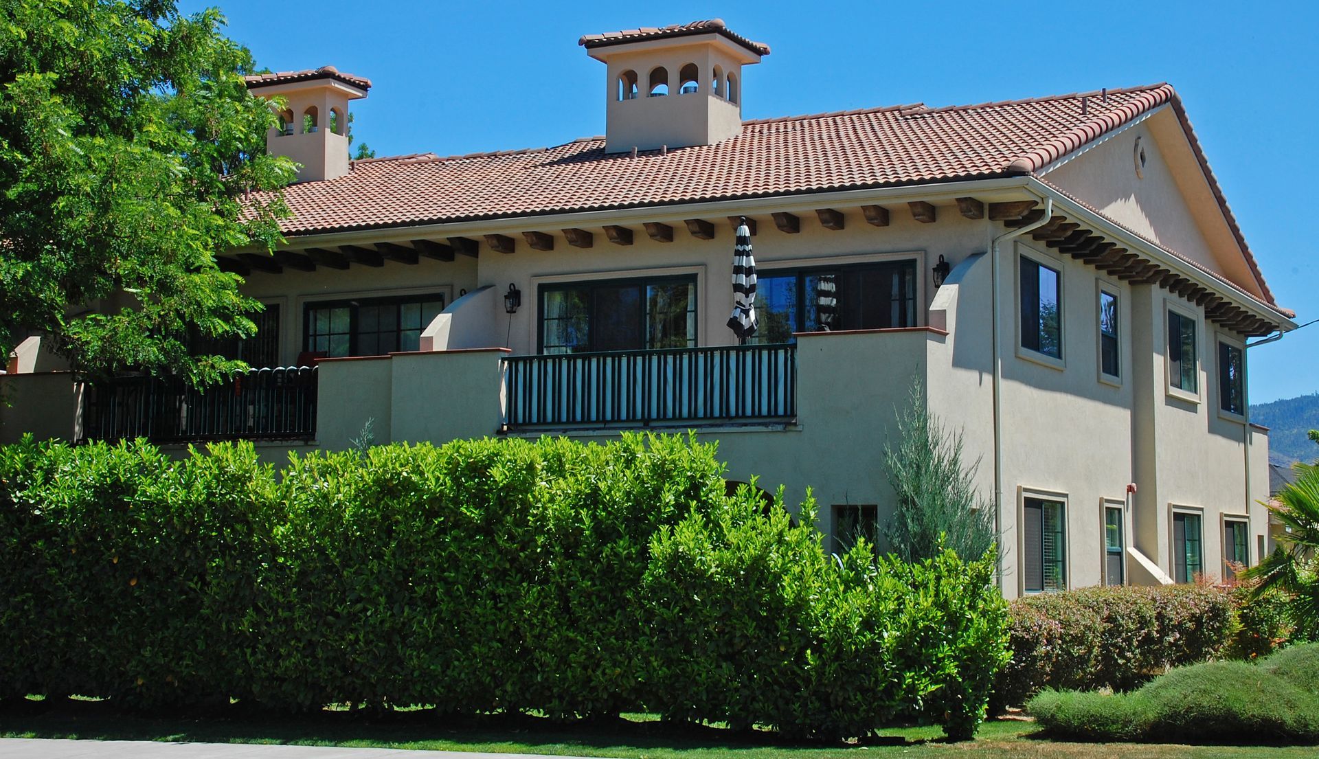 Spanish-style house with terracotta roof, blue balcony, and green hedges in bright sunlight