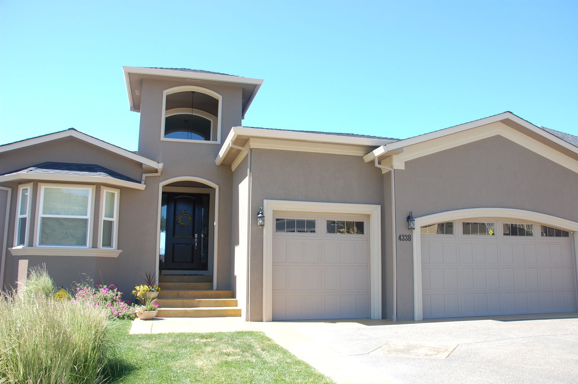 Single-story beige suburban house with two garage doors and a front porch under a clear blue sky