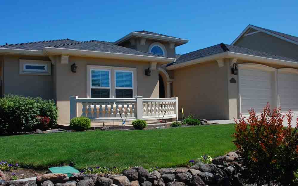 Single-story tan house with a small front porch, green lawn, and landscaped rocks under a blue sky