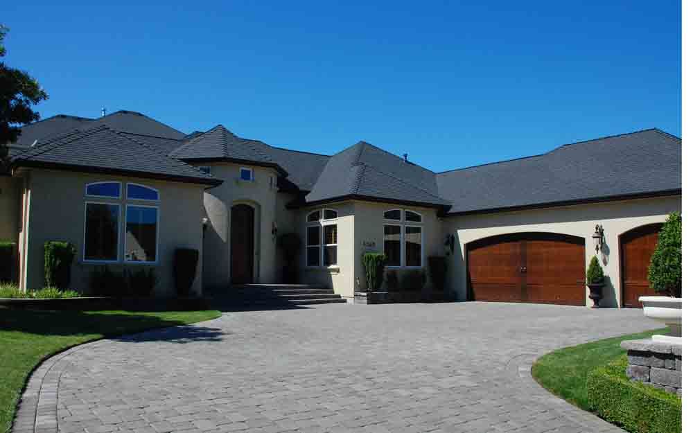 Large modern house with a curved driveway, gray roof, and wooden garage door under a clear blue sky