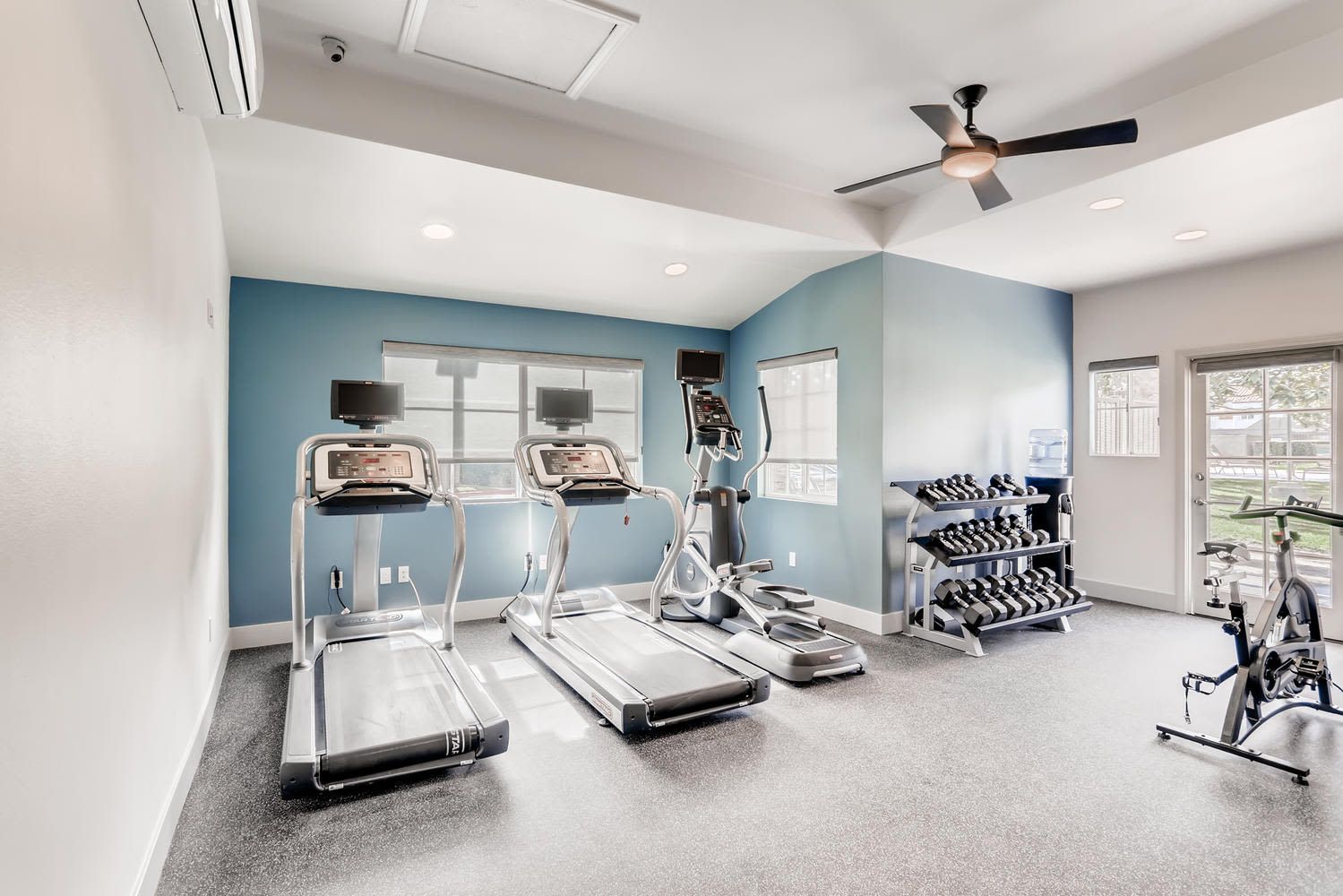 Well-lit community fitness room with treadmills, an elliptical, a dumbbell rack, and a water cooler.