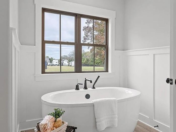 Oval bathtub beneath a window in a white bathroom with dark-framed window and fixtures, light gray walls.