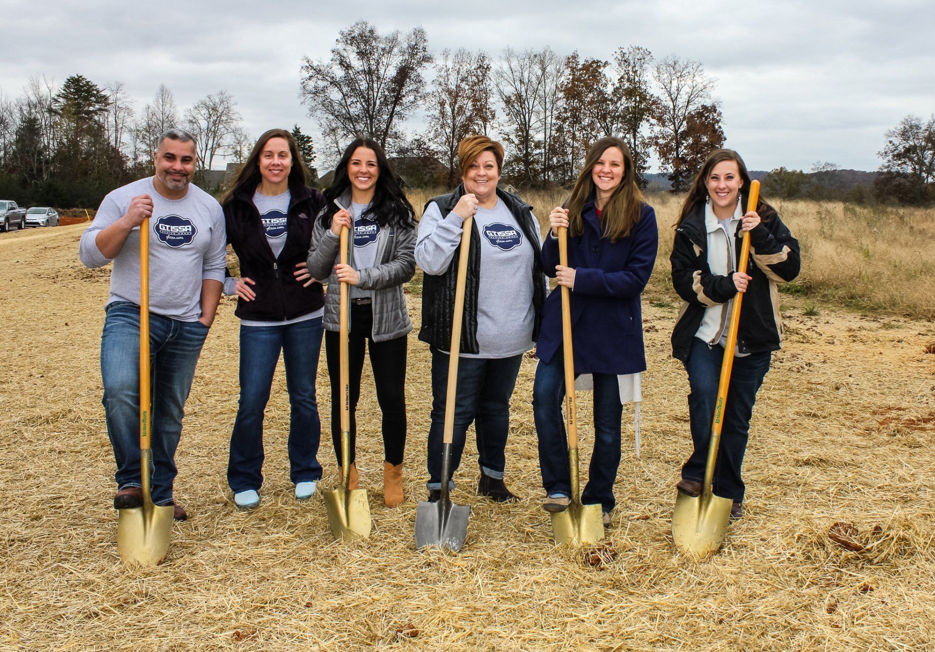 Six people holding shovels, standing in an open field, preparing for groundbreaking.