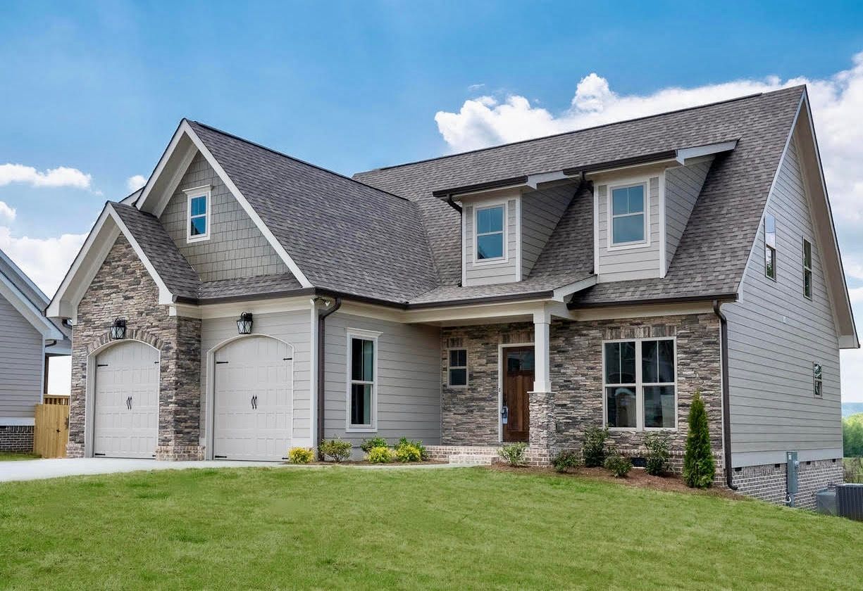 A two-story house with stone accents, a gray roof, and a green lawn under a blue sky.