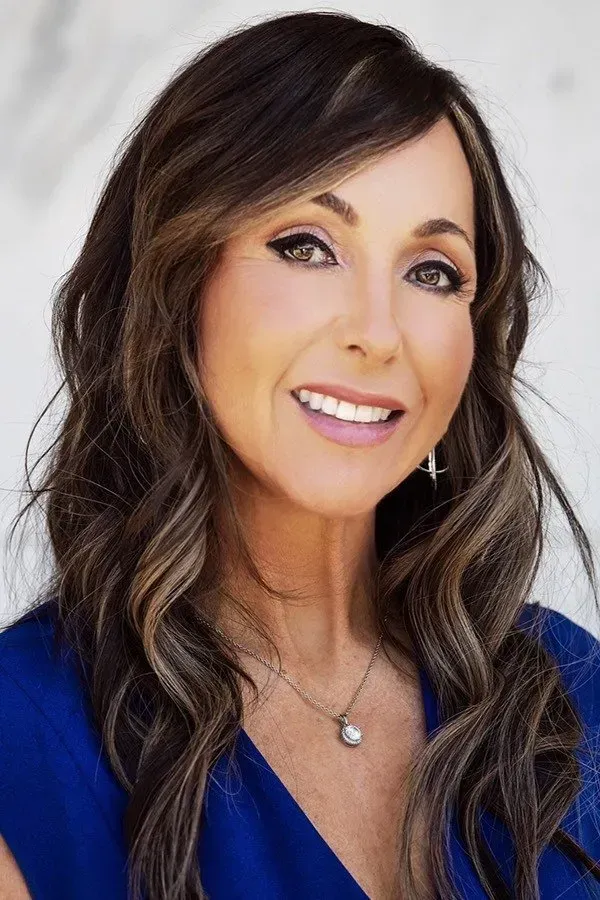 Woman with brown wavy hair in a blue top smiles, outdoors.