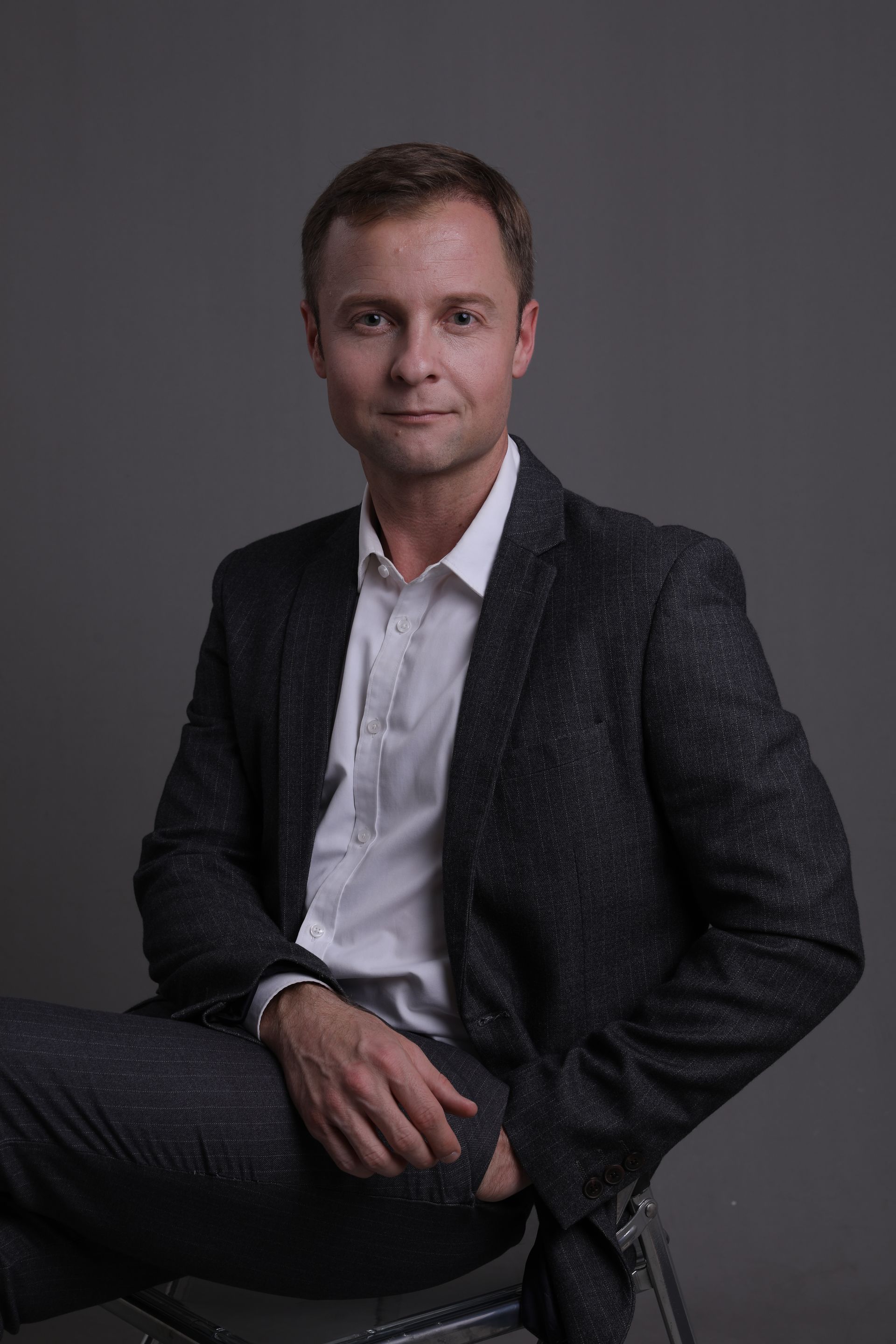 Seated professional in a dark blazer and white shirt against a gray studio backdrop