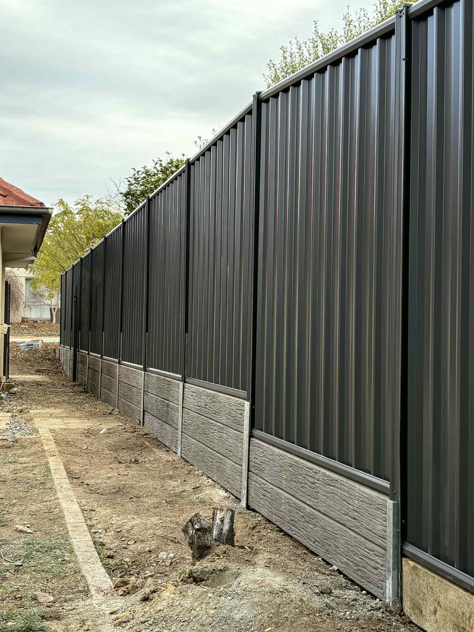 Long dark metal fence with stone base along a narrow dirt path beside a house. — GnB Fencing and Excavation in Queanbeyan, NSW