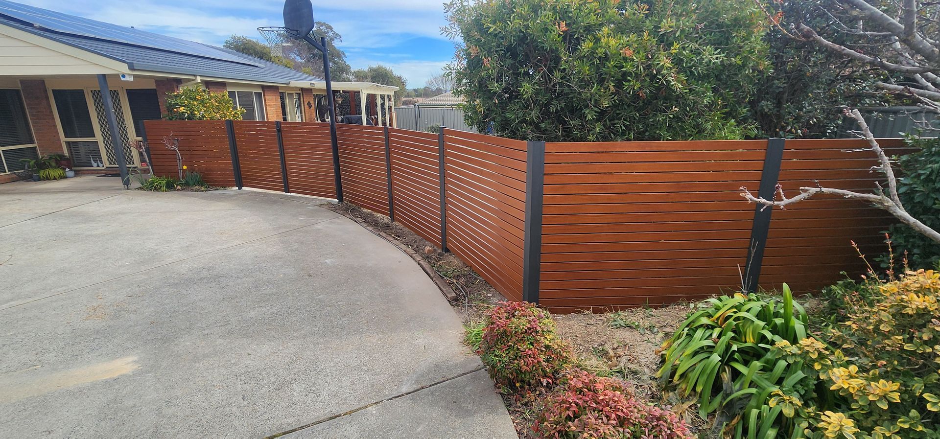 Curved wooden fence along a concrete driveway beside a garden under a sunny sky — GnB Fencing and Excavation in Queanbeyan, NSW