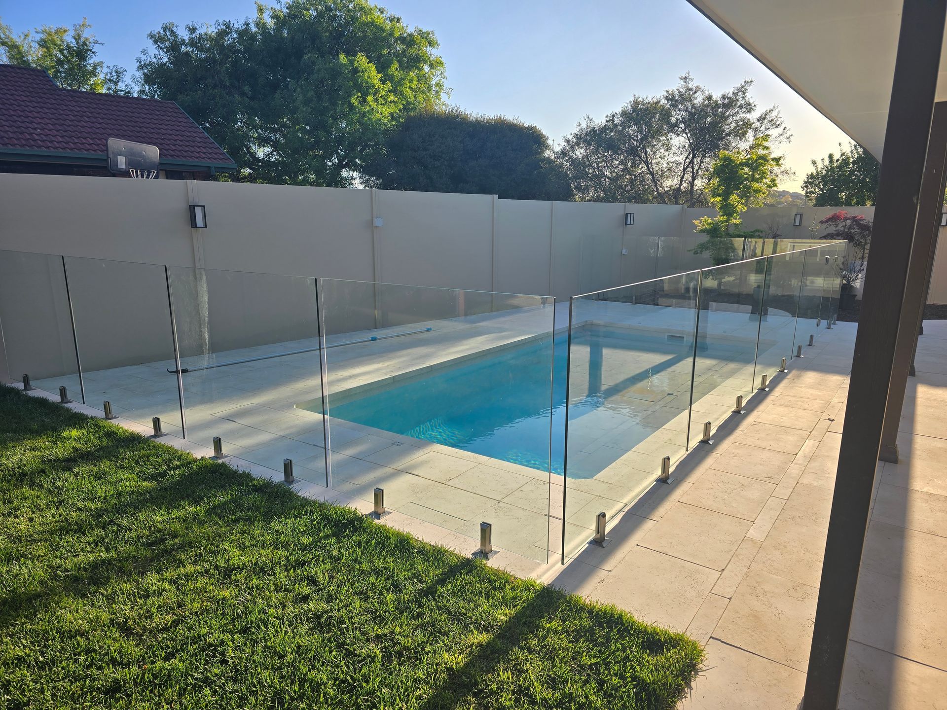 Backyard pool with glass fence, green lawn, and sunlit patio beside a white wall. — GnB Fencing and Excavation in Queanbeyan, NSW