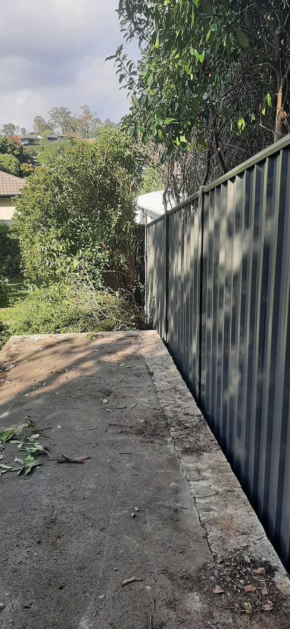 Narrow concrete path beside a tall gray metal fence, with trees and bushes in bright sunlight. — GnB Fencing and Excavation in Queanbeyan, NSW