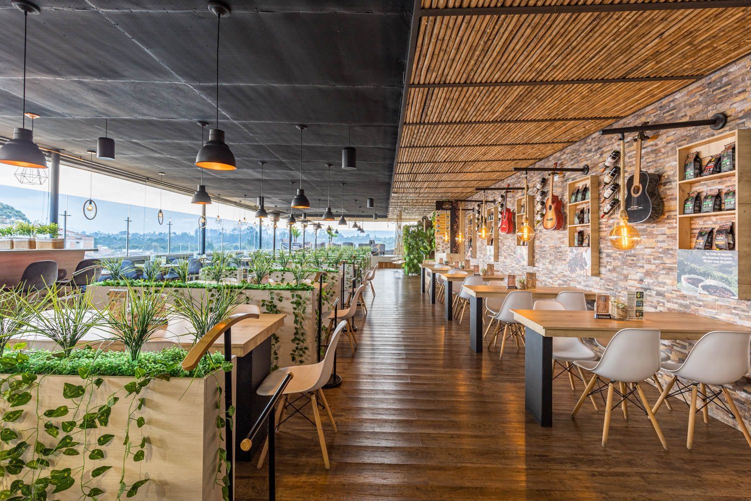 Interior del restaurante: la mitad con plantas y techo negro, la otra mitad con techo de bambú y pared de ladrillo con guitarras. Suelo de madera y sillas blancas.