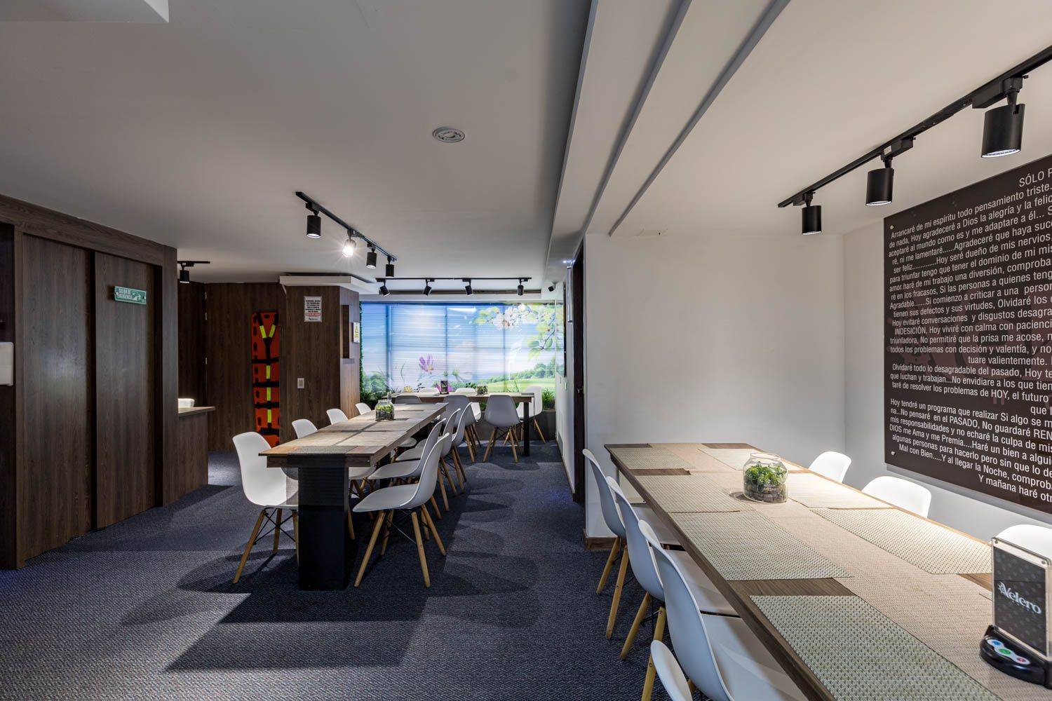 Un comedor largo y moderno con varias mesas, sillas e iluminación en riel. Una gran obra de arte cuelga de la pared.