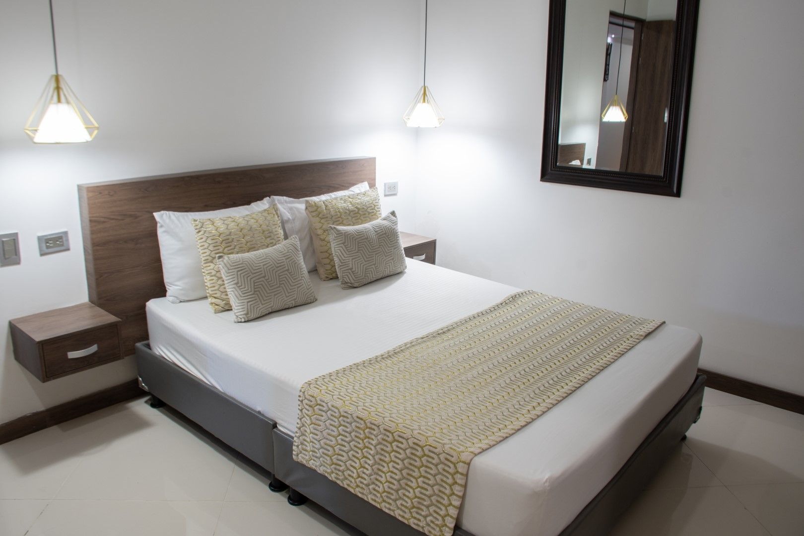 Dormitorio con cama blanca, cabecero de madera, mesitas de noche y cojines decorativos. Hay un espejo en la pared y lámparas colgantes.