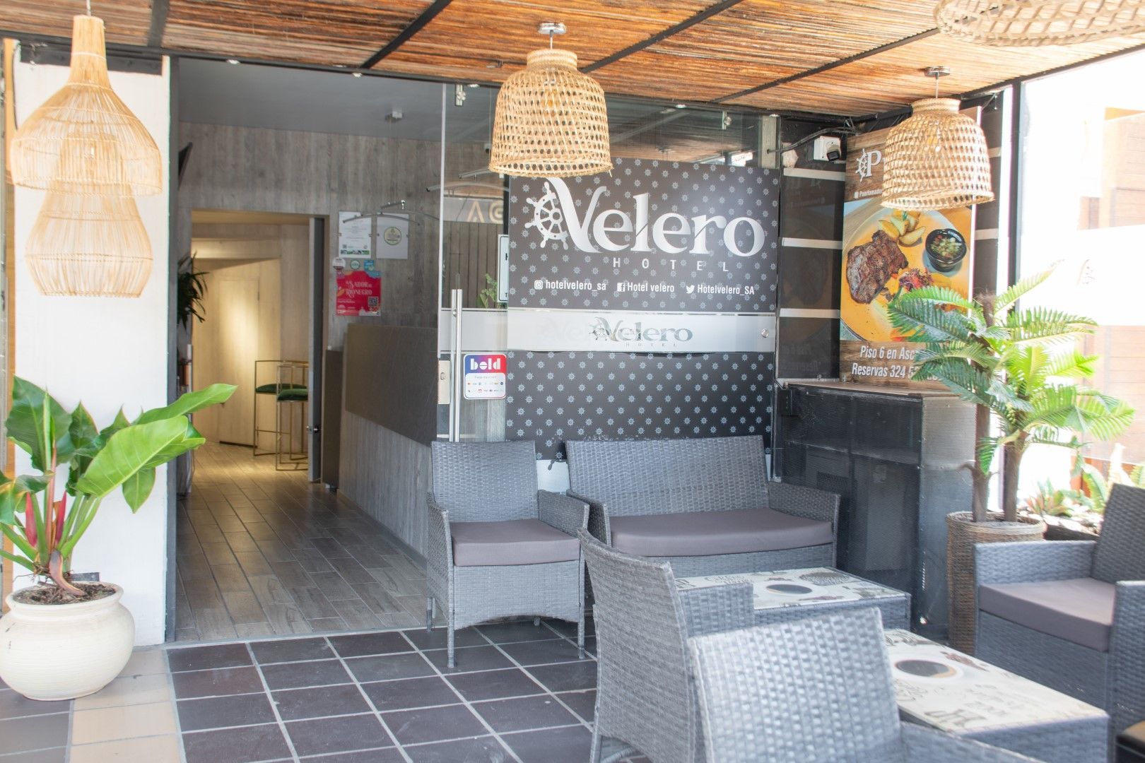 Entrada al Restaurante Velero, con lámparas colgantes de mimbre, asientos al aire libre y puertas de vidrio con el logotipo del restaurante.