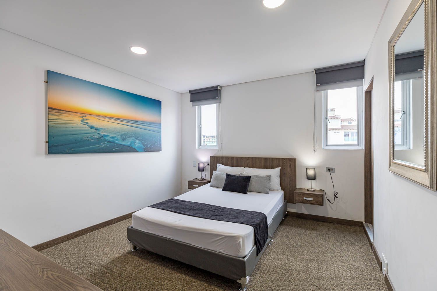 Dormitorio con cama de matrimonio, colores neutros, obra de arte sobre la cama y dos pequeñas ventanas con cortinas.