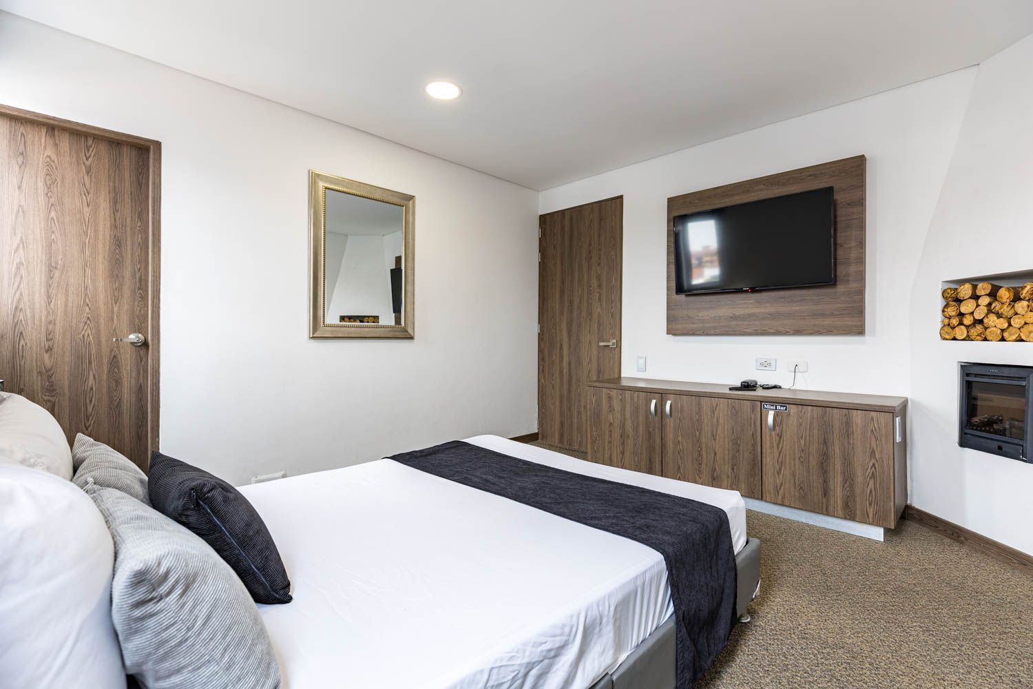 Un dormitorio moderno con cama, televisor, chimenea y detalles en madera. La habitación está muy iluminada, con paredes blancas y alfombra.