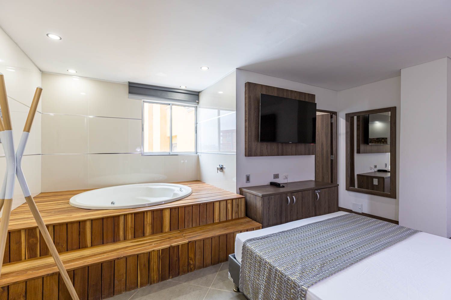 Una moderna habitación de hotel con una plataforma elevada de madera que alberga un jacuzzi. Un televisor y un espejo están colgados en la pared.