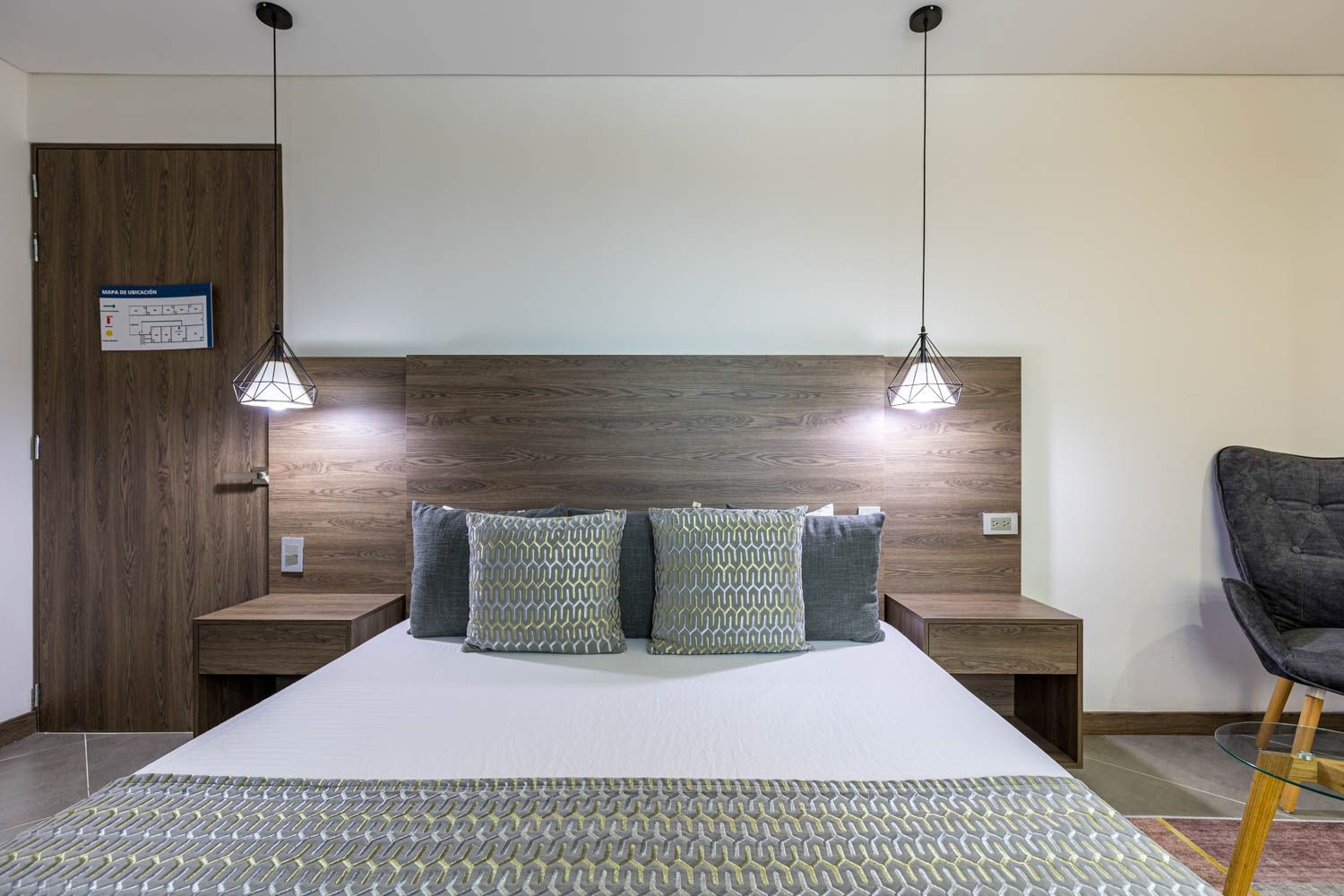 Dormitorio con una cama, almohadas, dos mesitas de noche, lámparas colgantes, una puerta y un sillón gris.