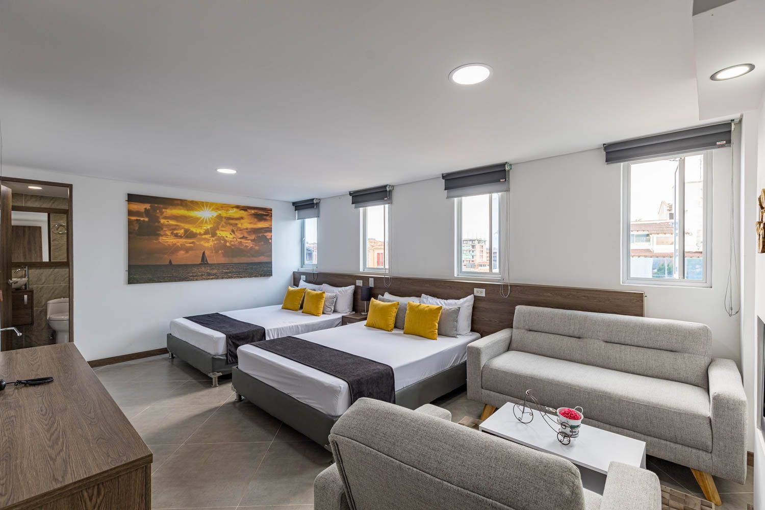 Una habitación de hotel con dos camas dobles, un sofá y un cuadro de una puesta de sol. Paredes claras, ventanas y muebles en tonos neutros.