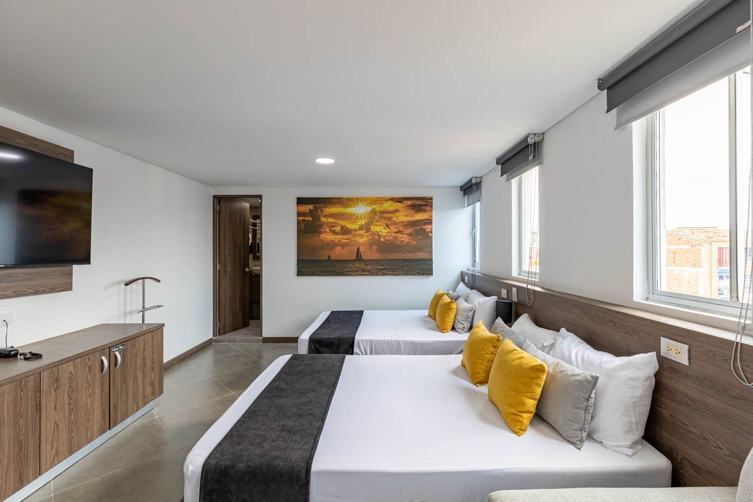 Amplia habitación de hotel con dos camas dobles, paredes blancas, un gran cuadro y un televisor colgado.
