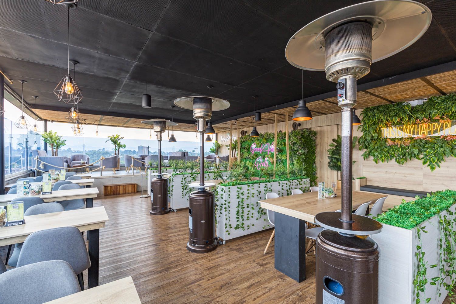 Restaurante en la azotea con estufas de exterior, mesas y vegetación. Suelos de madera, techo negro y vistas a la ciudad.