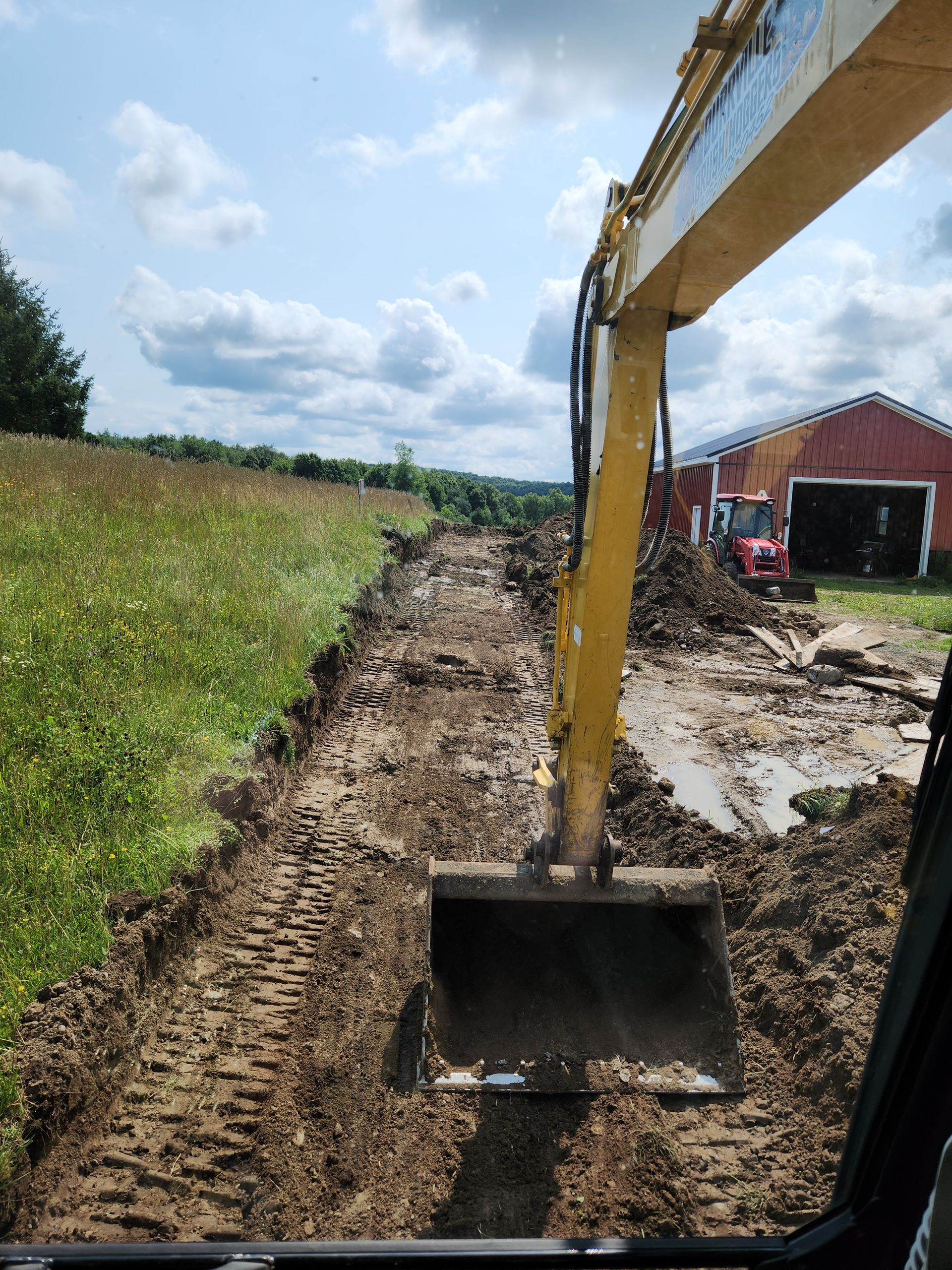Yellow excavator digging a muddy trench beside a grassy field and red barn under a blue sky