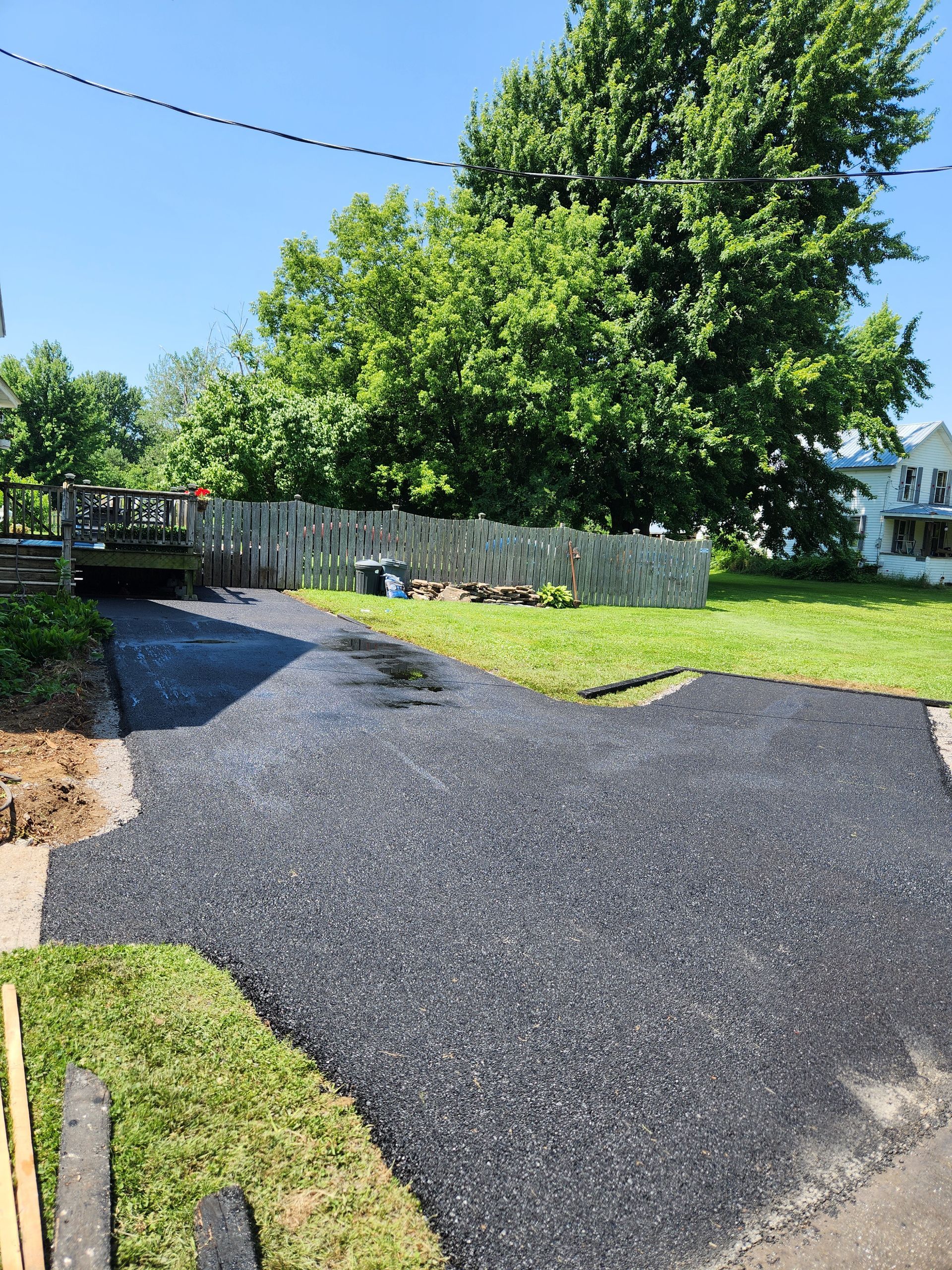 Fresh black asphalt driveway curving through a green yard beside a wooden fence and large tree