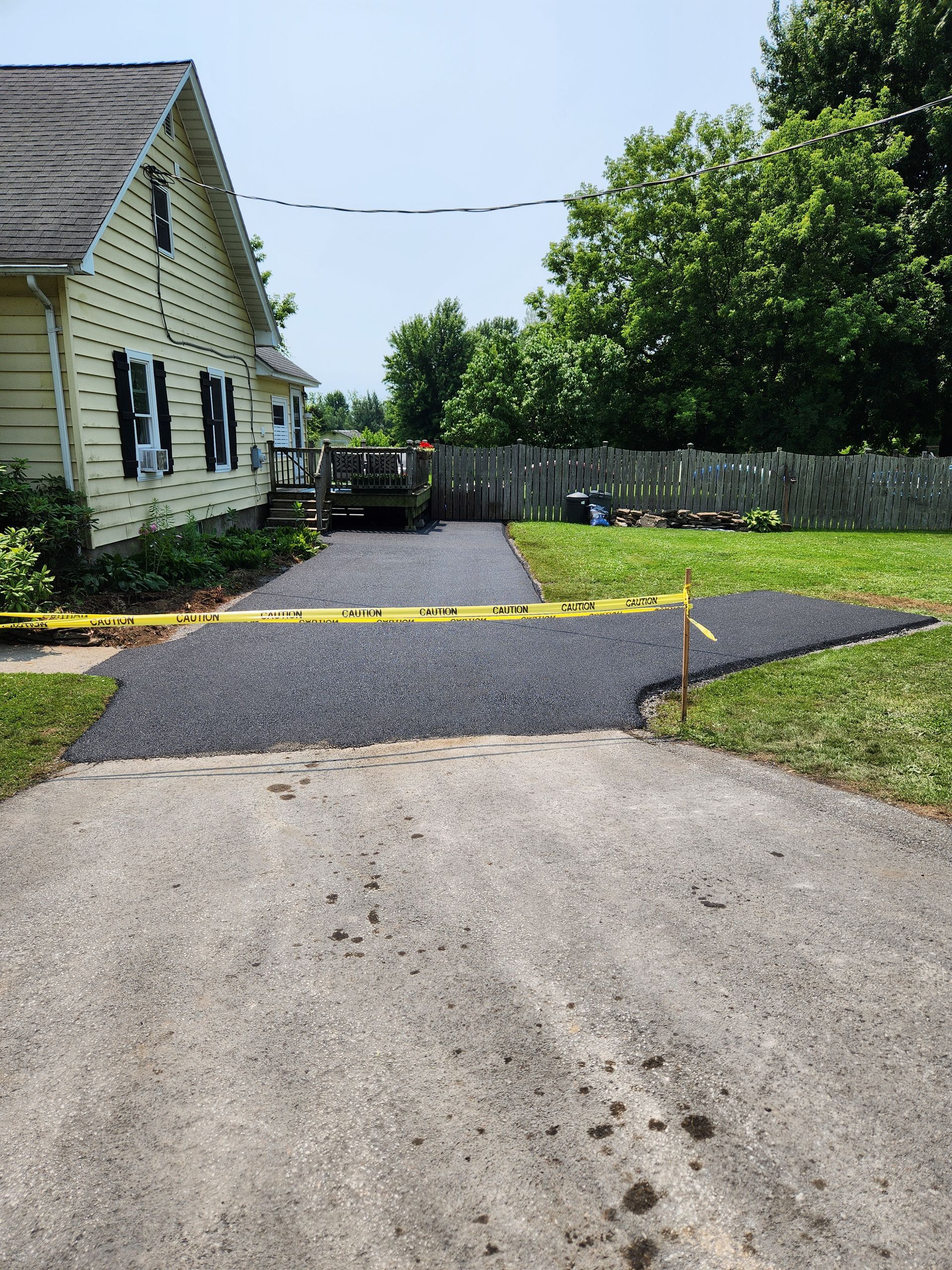 Driveway with a yellow speed bump, house on the left, trees and a fence in a sunny backyard.