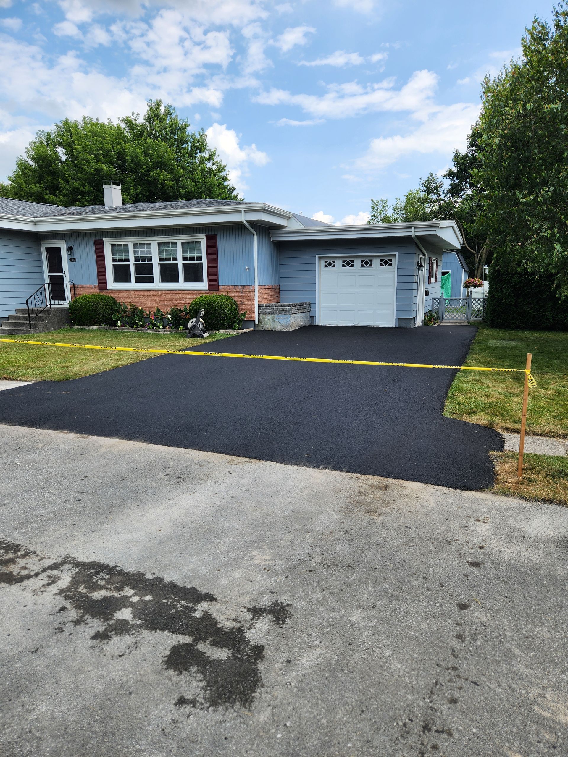 Single-story blue house with new black driveway, attached garage, and green trees under a partly cloudy sky