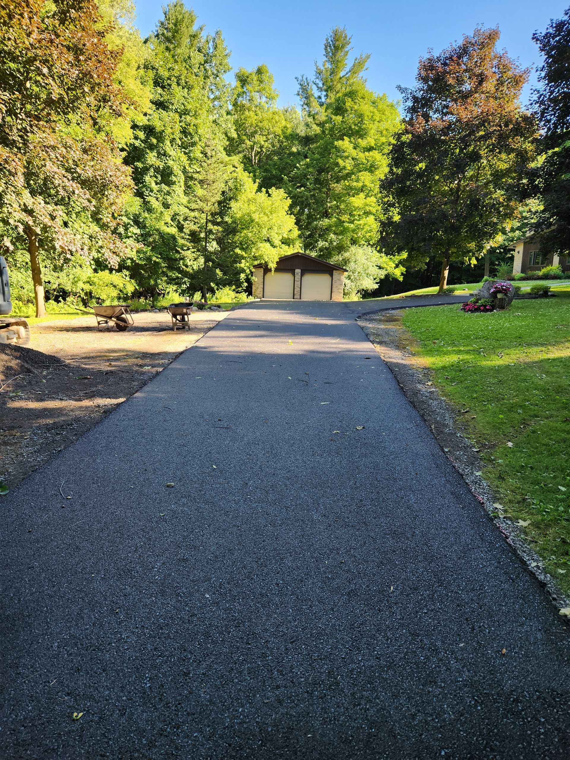 Gravel driveway leading to a small garage, surrounded by green trees and a sunny yard