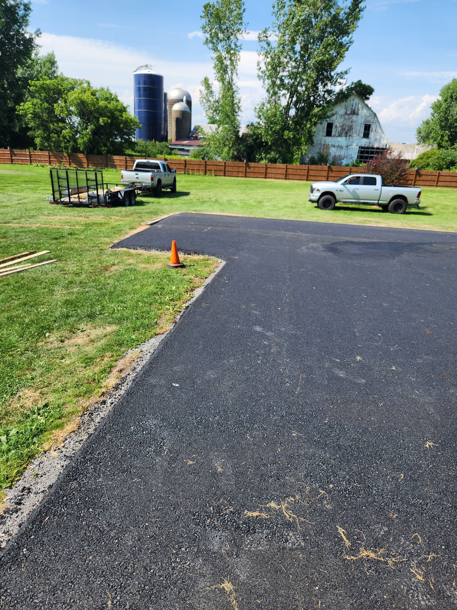 Gravel lot with parked cars, orange cone, grassy field, and farm buildings under a blue sky