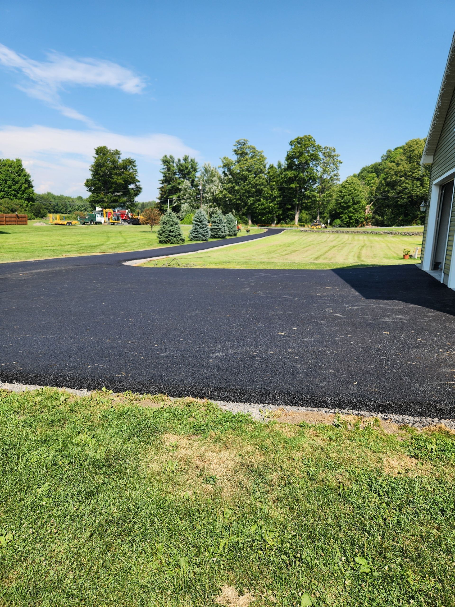 Freshly paved black driveway beside a grassy yard with trees under a blue sky