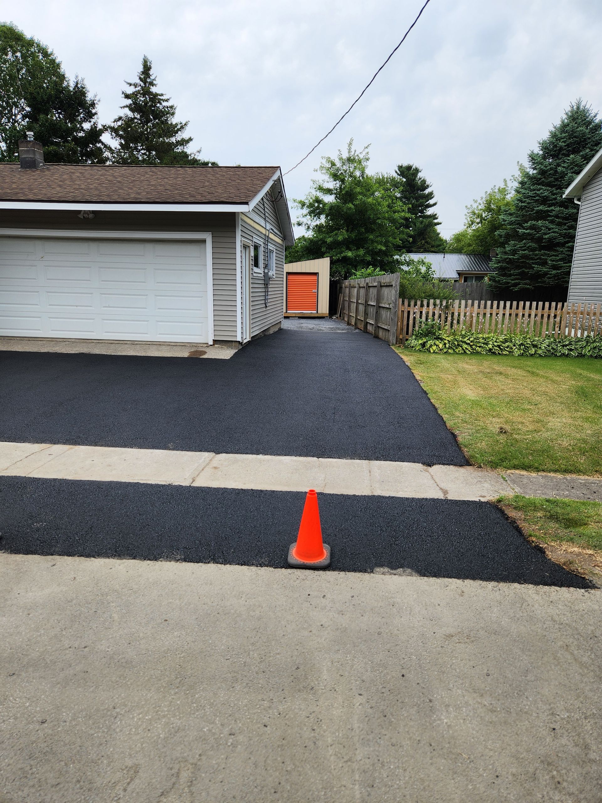 Driveway with orange cone, black asphalt, garage, and fenced backyard under cloudy sky.