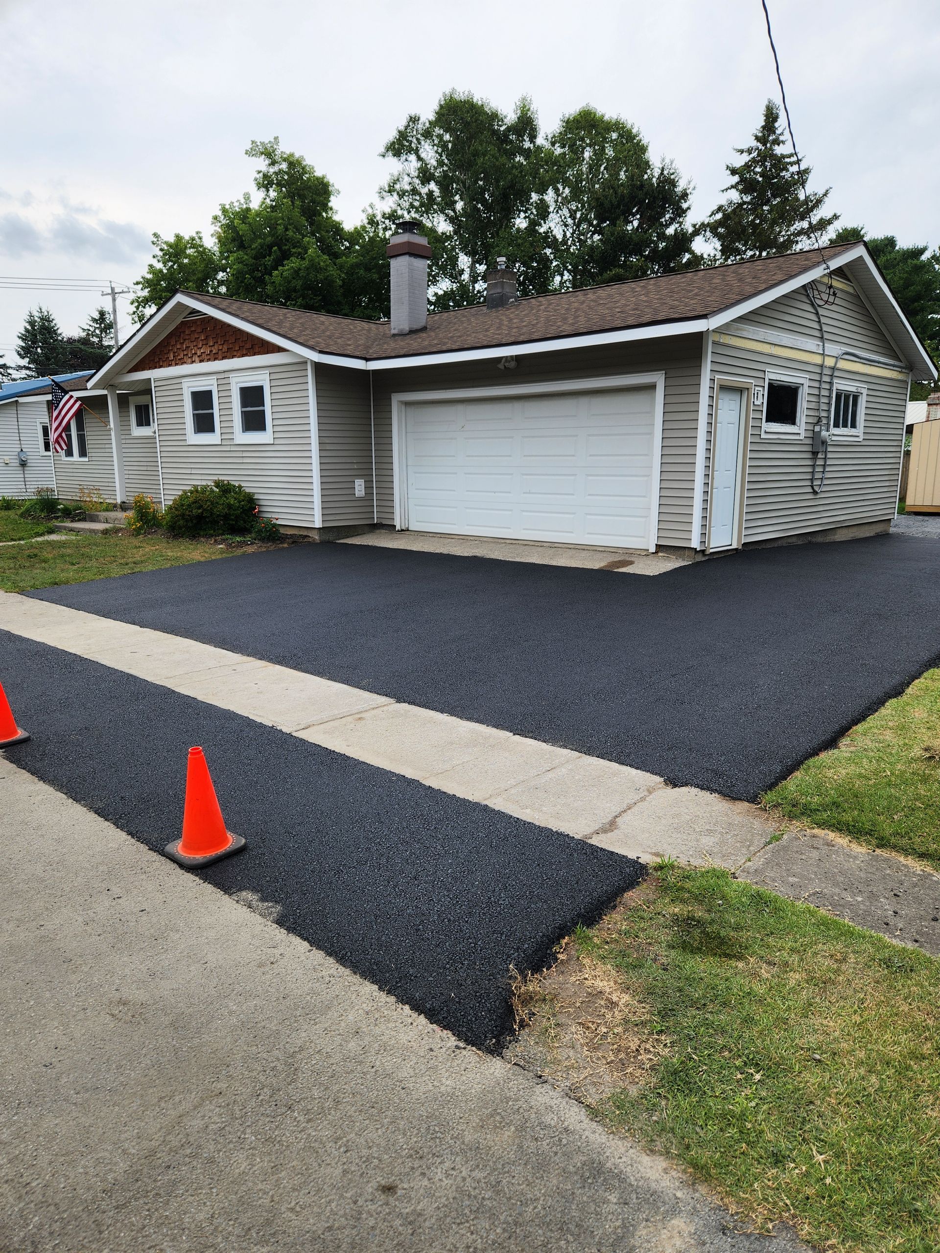 White house with garage and fresh black driveway paving, marked by orange cones under cloudy skies