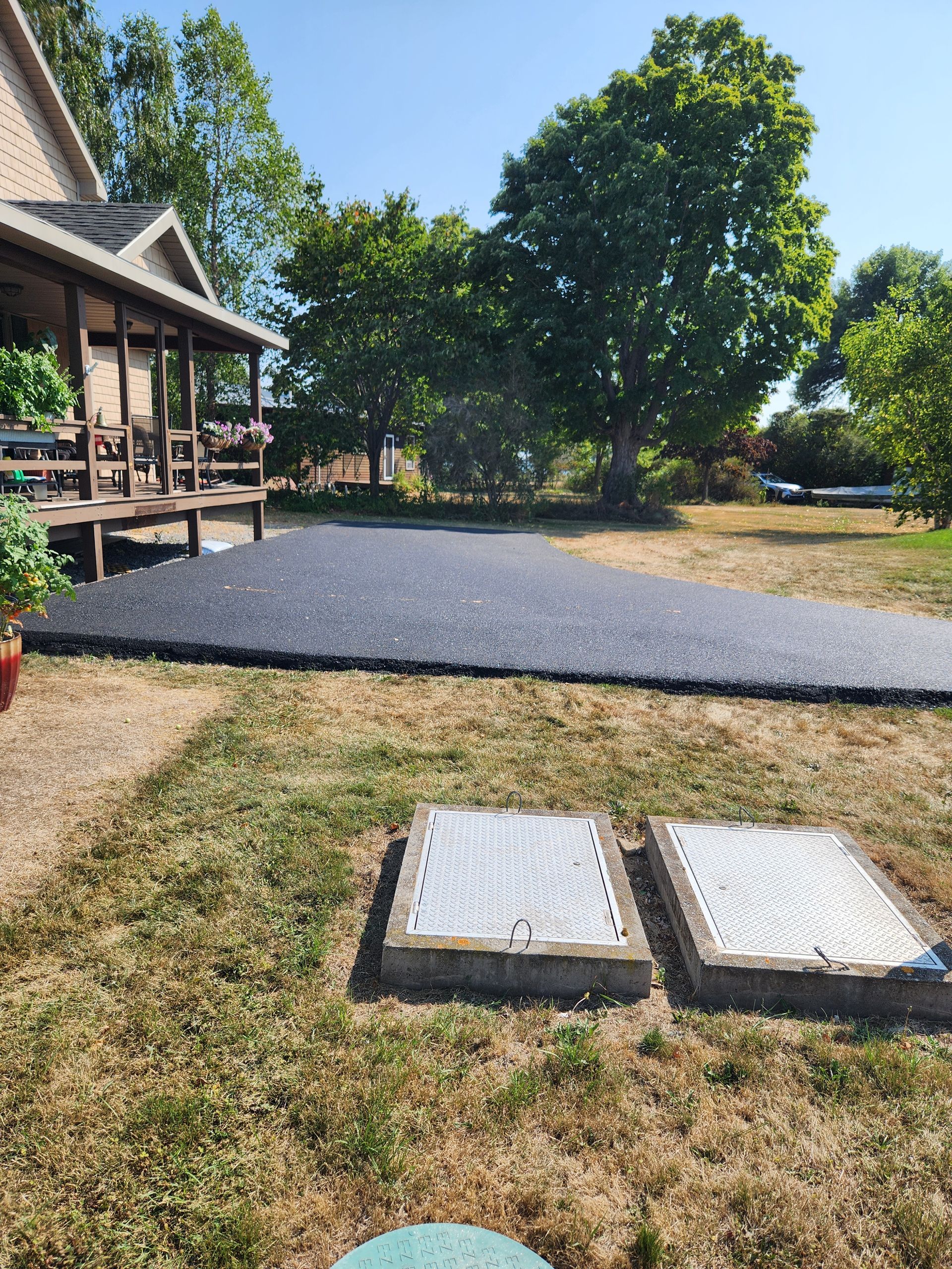 Porch beside a gravel patio and two utility covers in a dry grassy yard with trees and a house.
