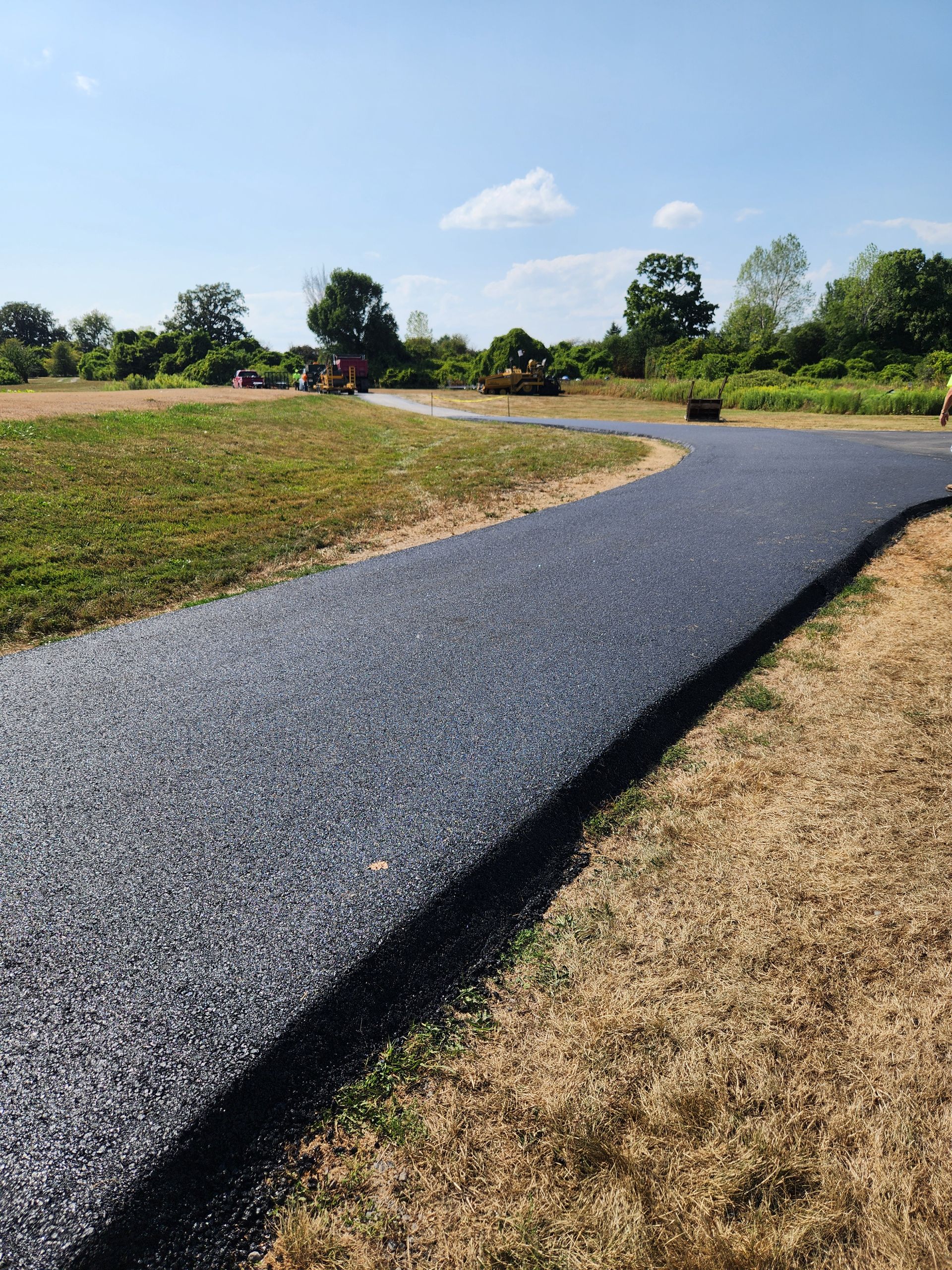 Winding black gravel path through a sunny grassy park with trees and blue sky