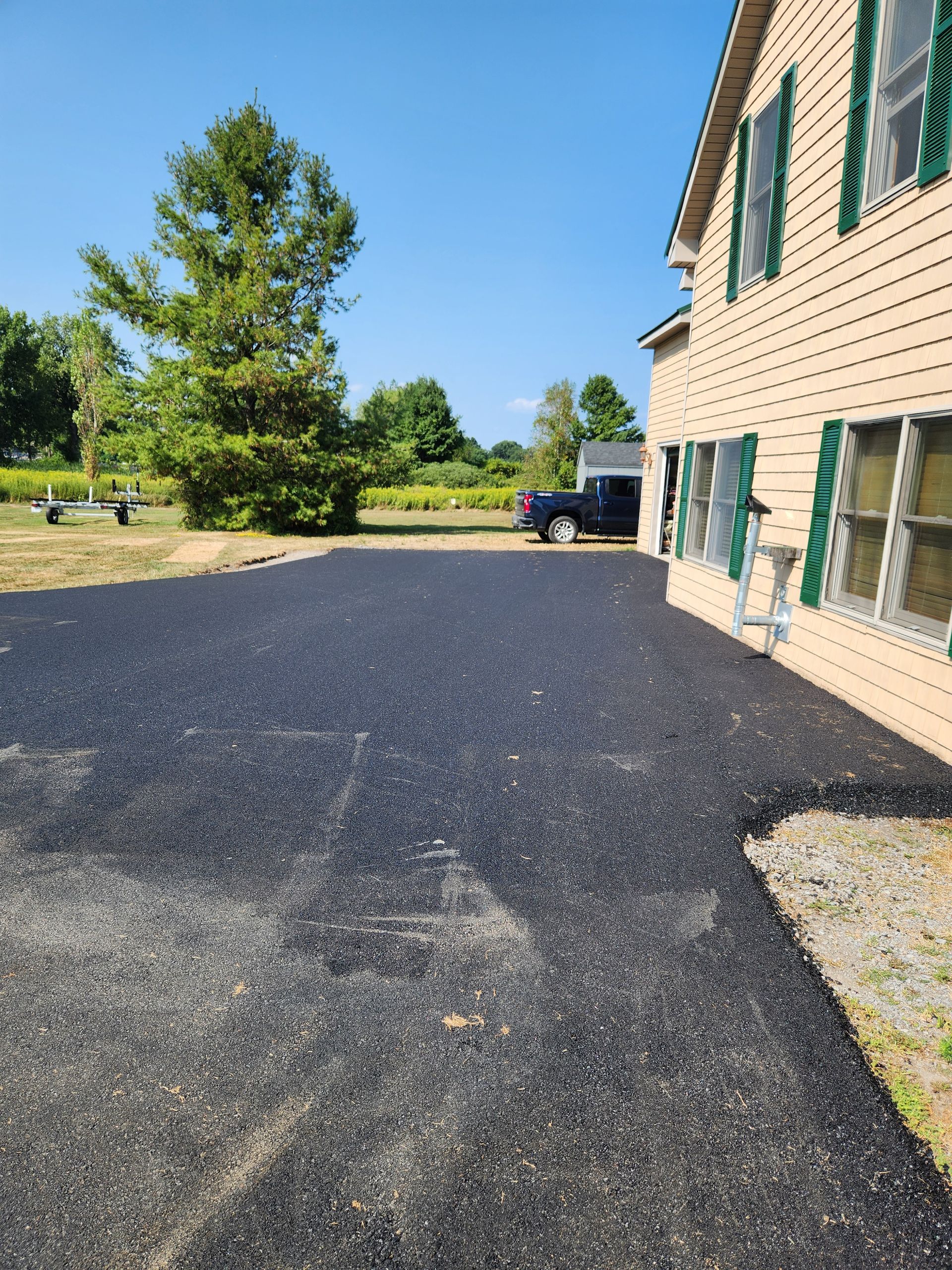 Gravel driveway beside a beige house with green shutters, trees, and a parked truck under a clear blue sky