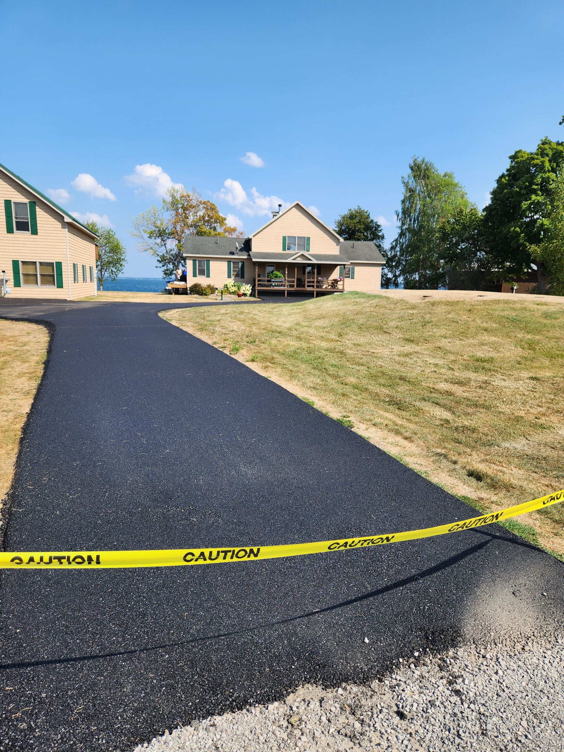 Yellow caution tape across a black paved driveway leading to a house on a sunny suburban lot