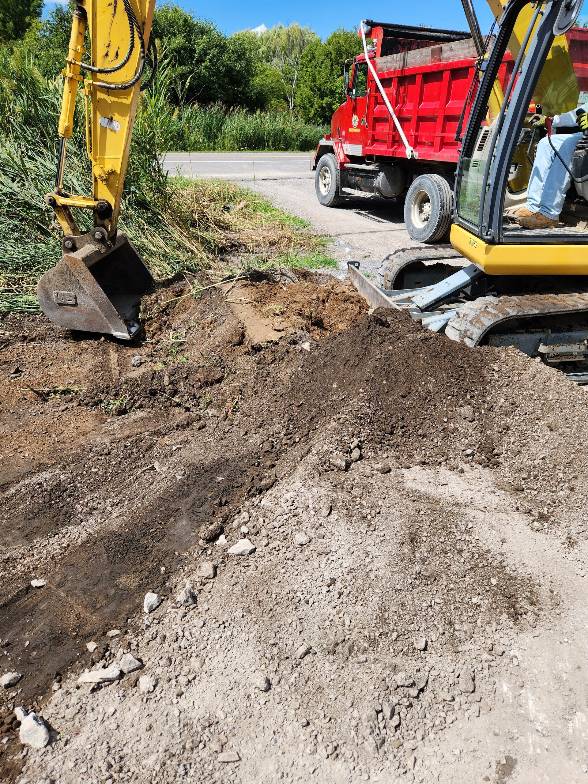 Excavator digging soil beside a red dump truck on a roadside construction site