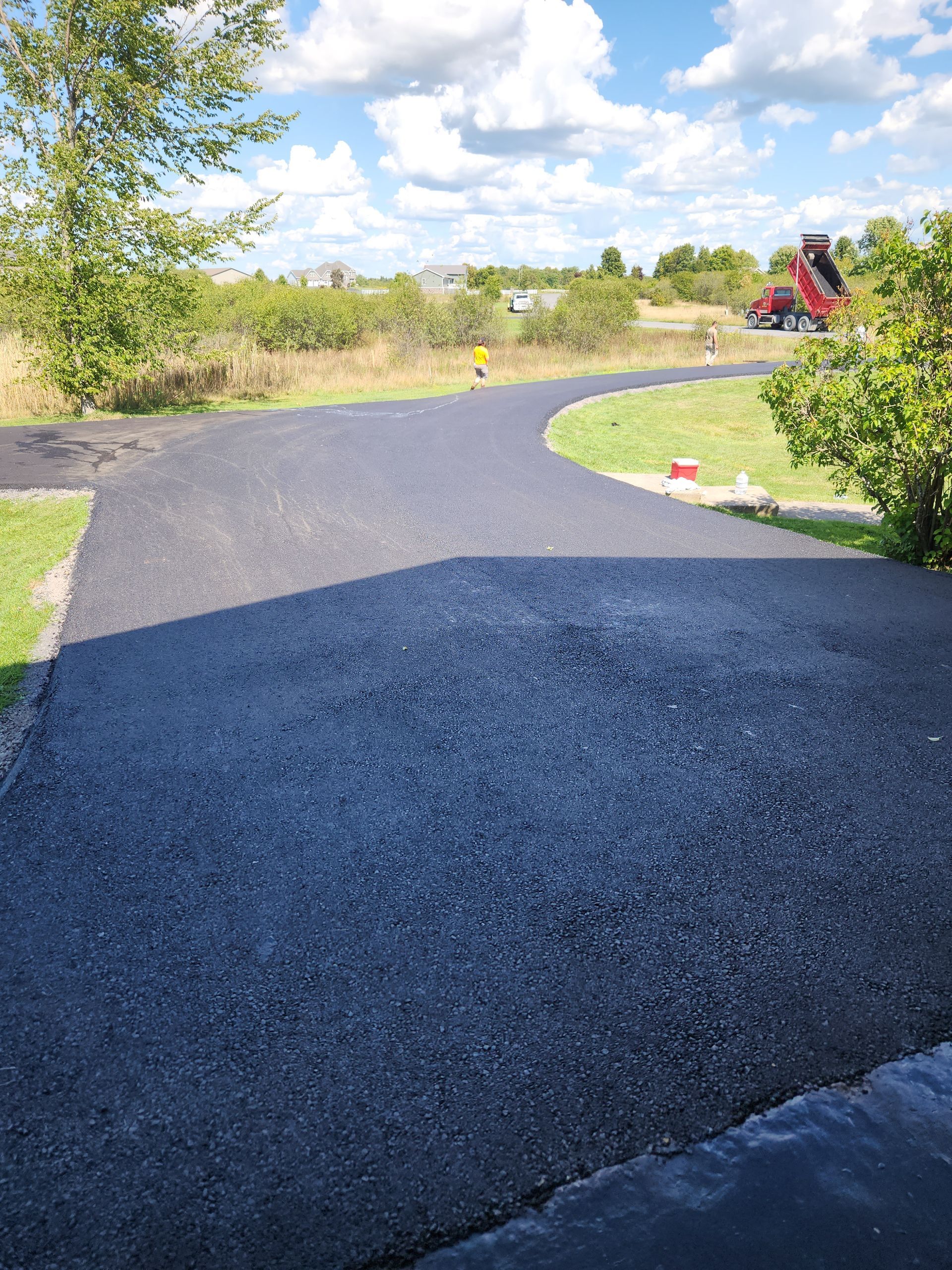 Curving gravel driveway beside trees and a grassy field under a bright blue sky