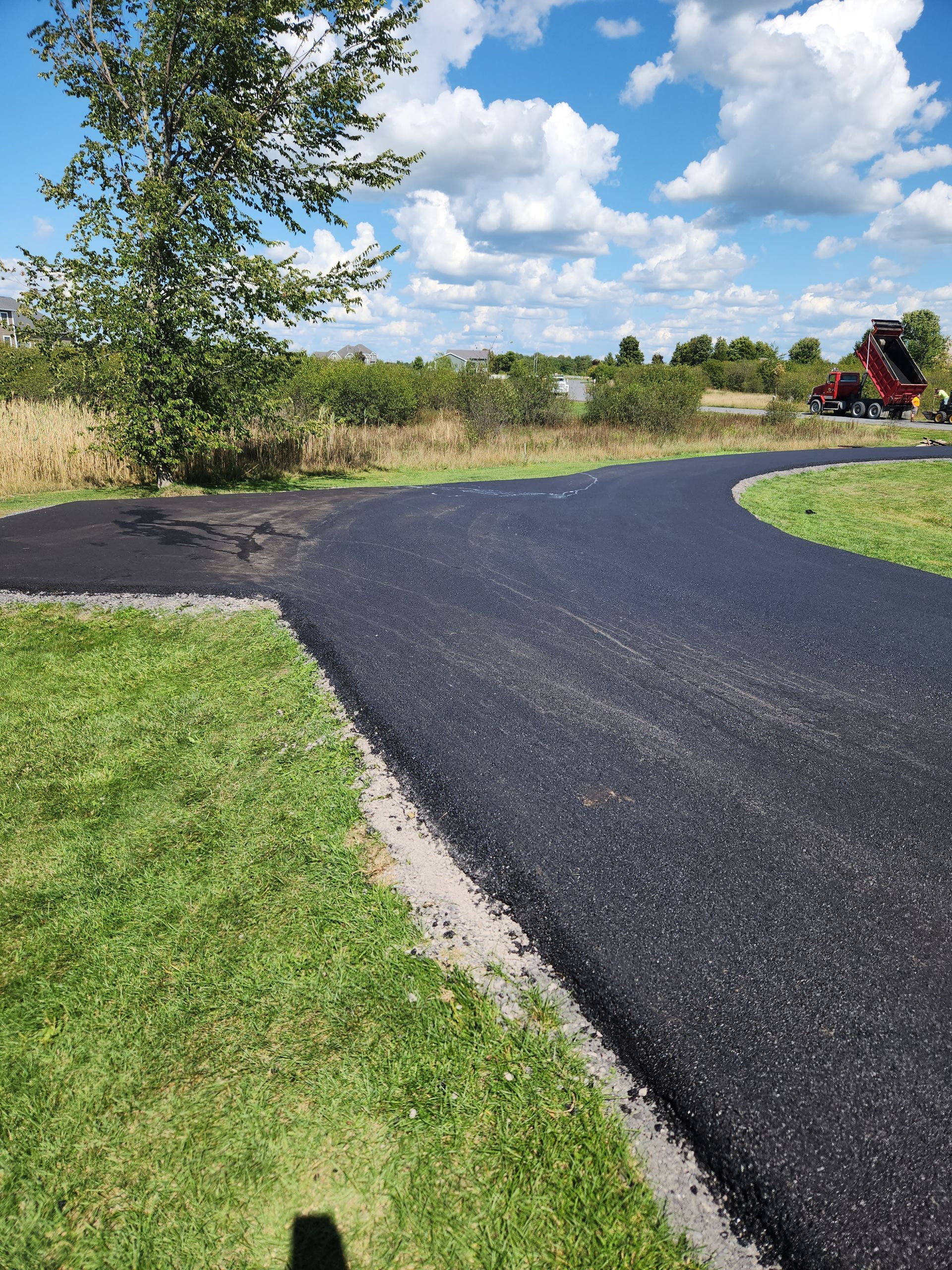 Curved black asphalt path through a grassy field under a blue sky with clouds, a tree at left, and a tractor in the distance