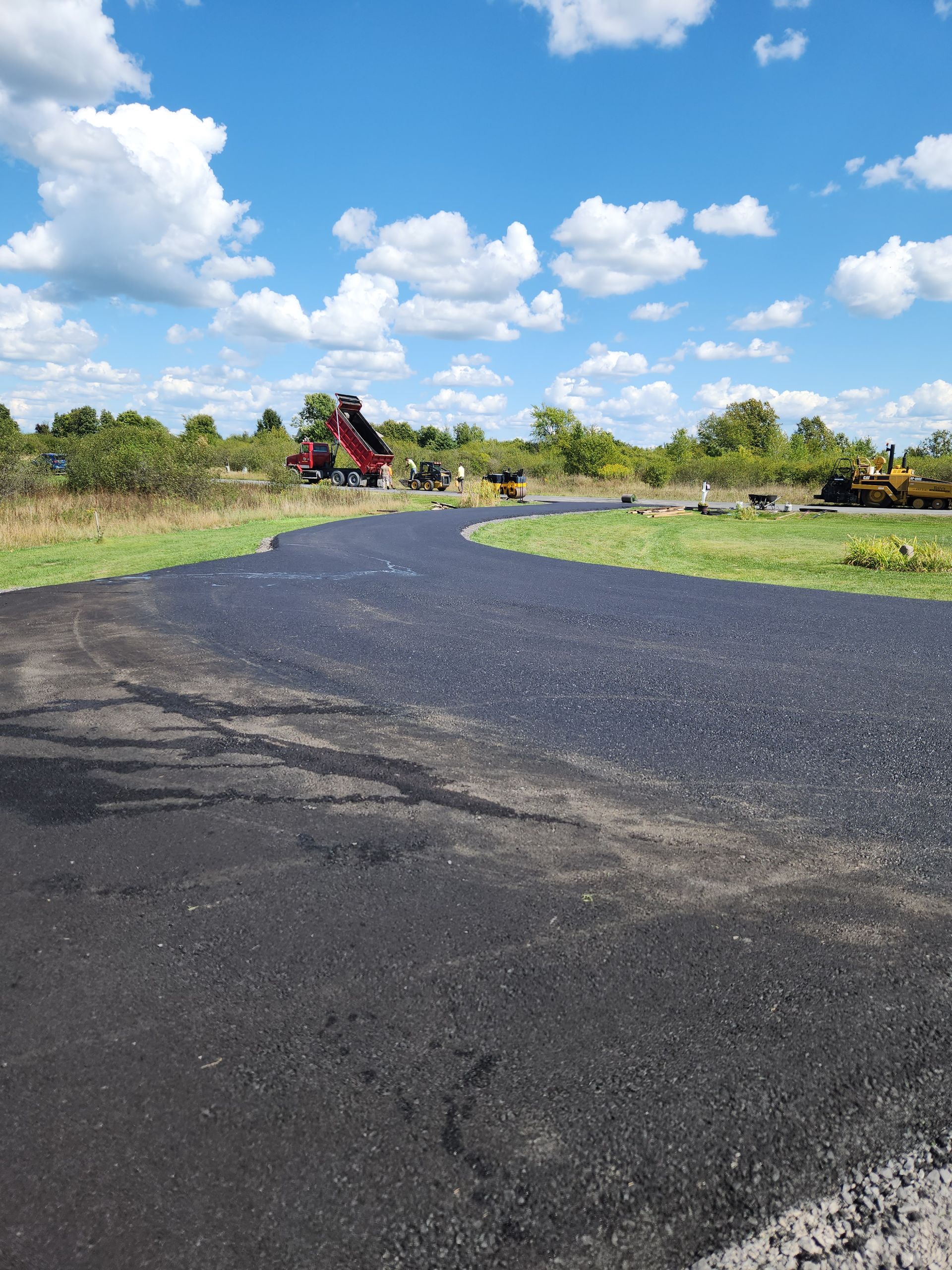 Curving black gravel road through green fields under a bright blue sky with clouds and a distant red tractor