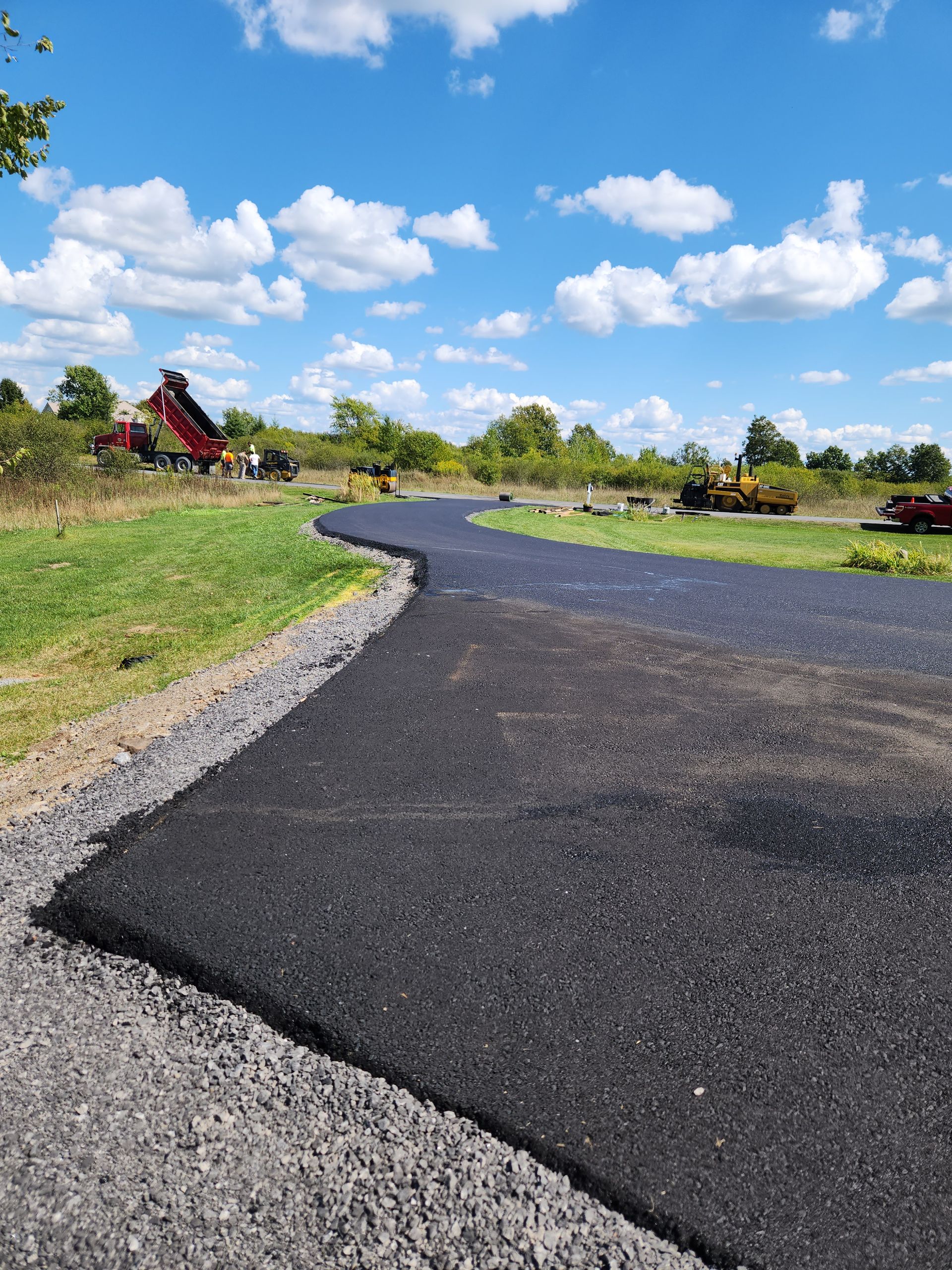 Fresh black asphalt path winding through a grassy landscape under a blue sky with clouds