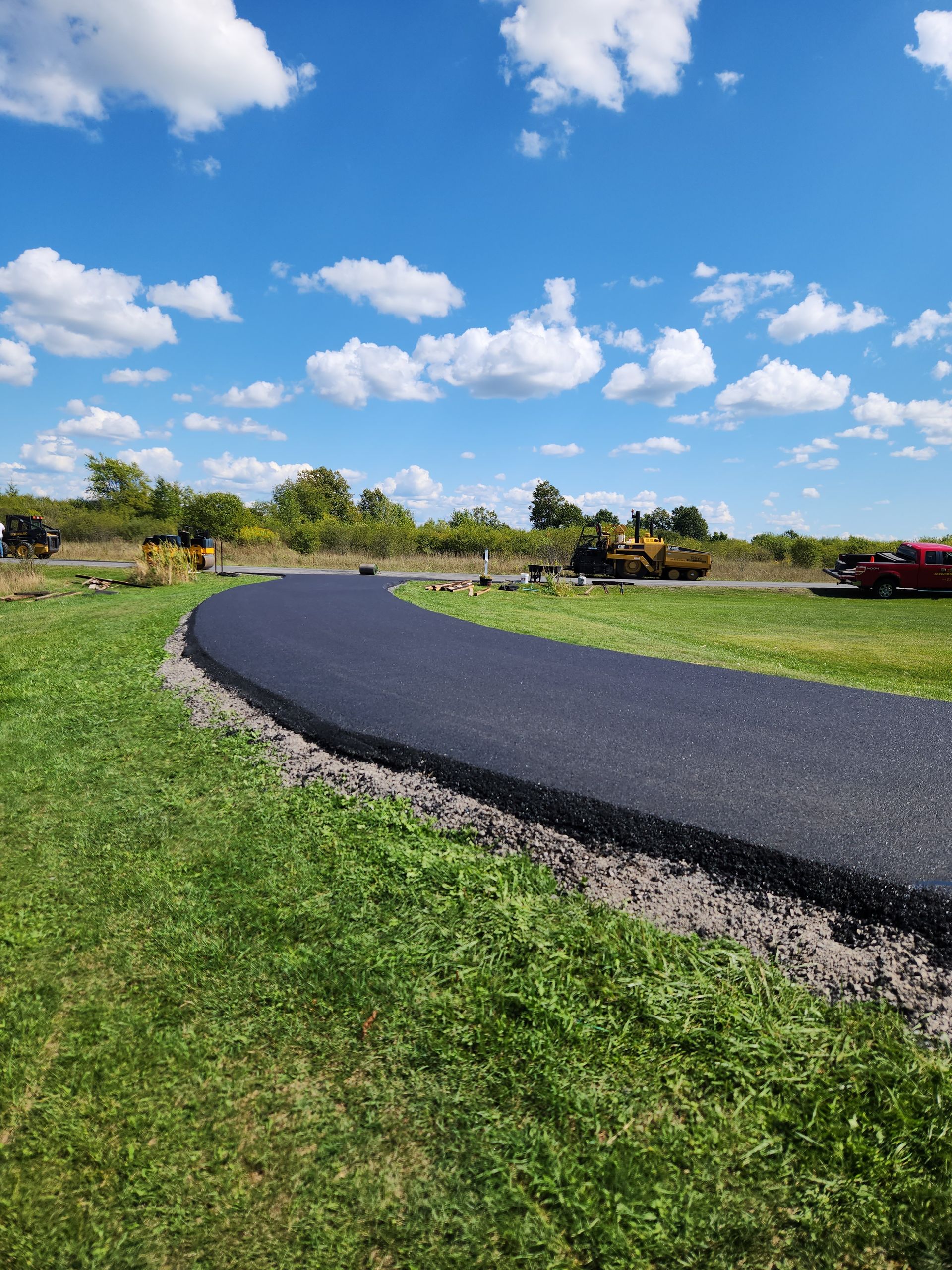 Curved black asphalt road under construction across a grassy field, with machinery and a cloudy blue sky.