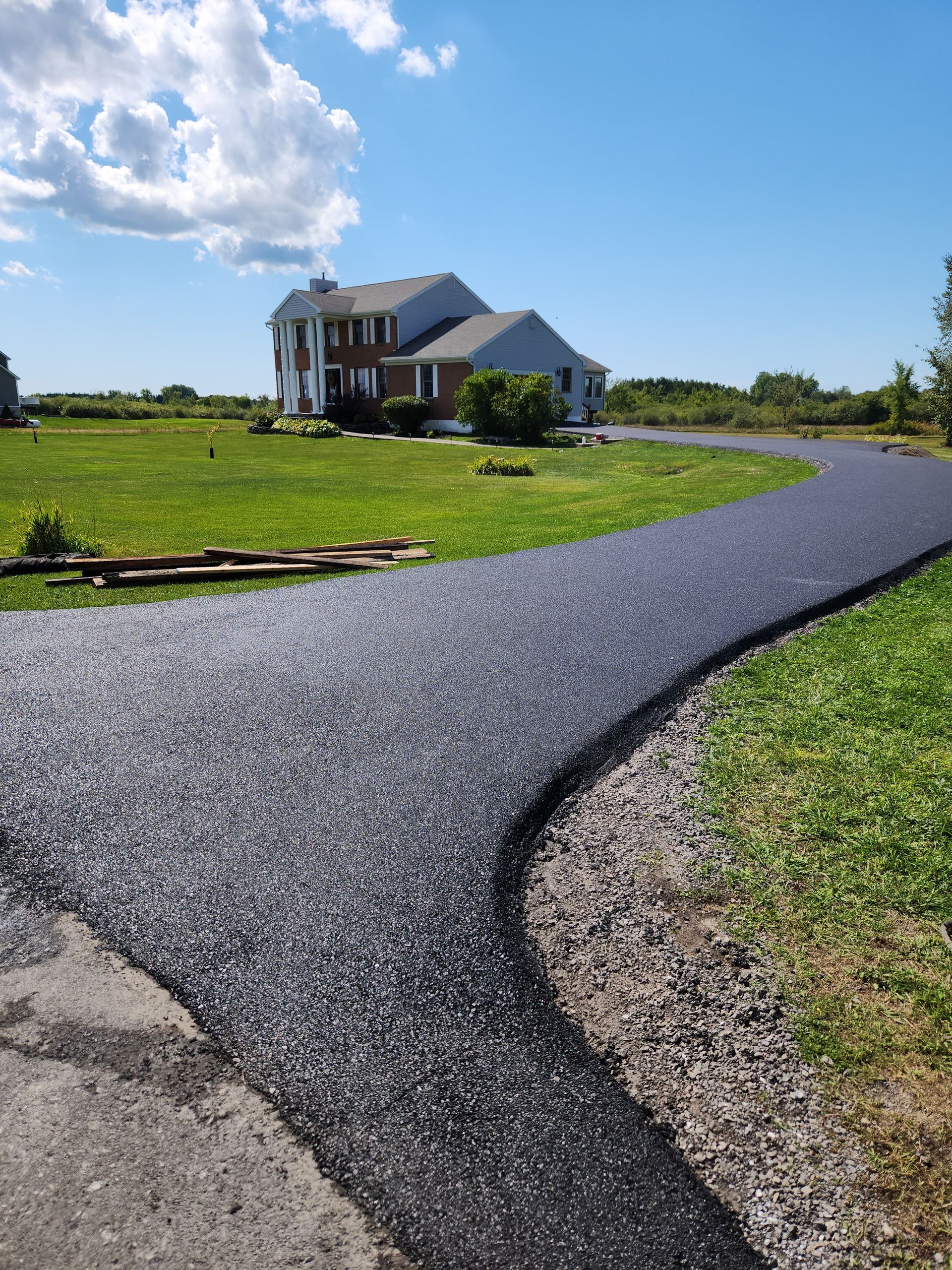 Curved gravel driveway leading to a house on a sunny green lawn under a blue sky