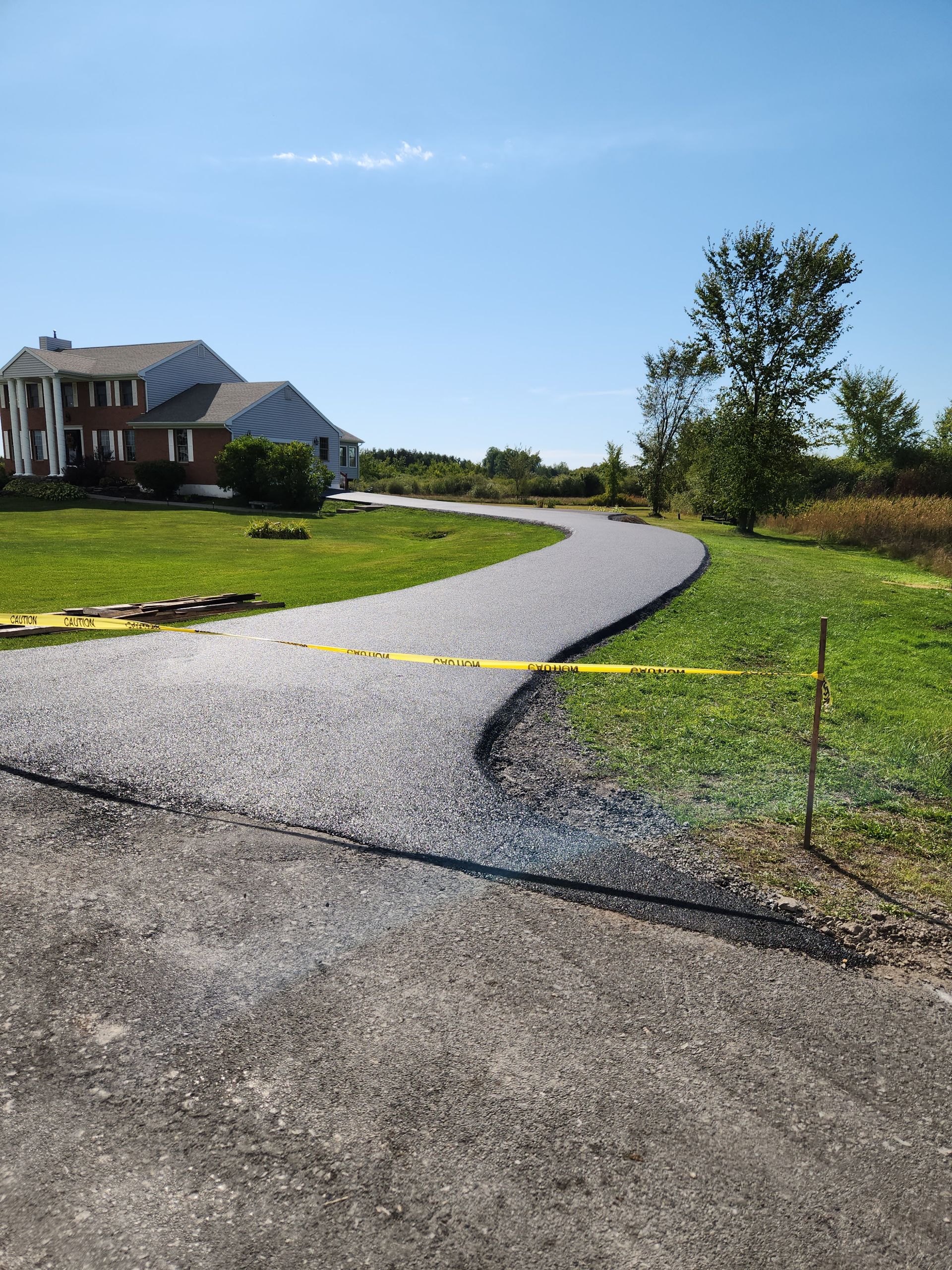Curving gravel driveway leading to a large house, with yellow caution tape and a tree-lined lawn under a blue sky.